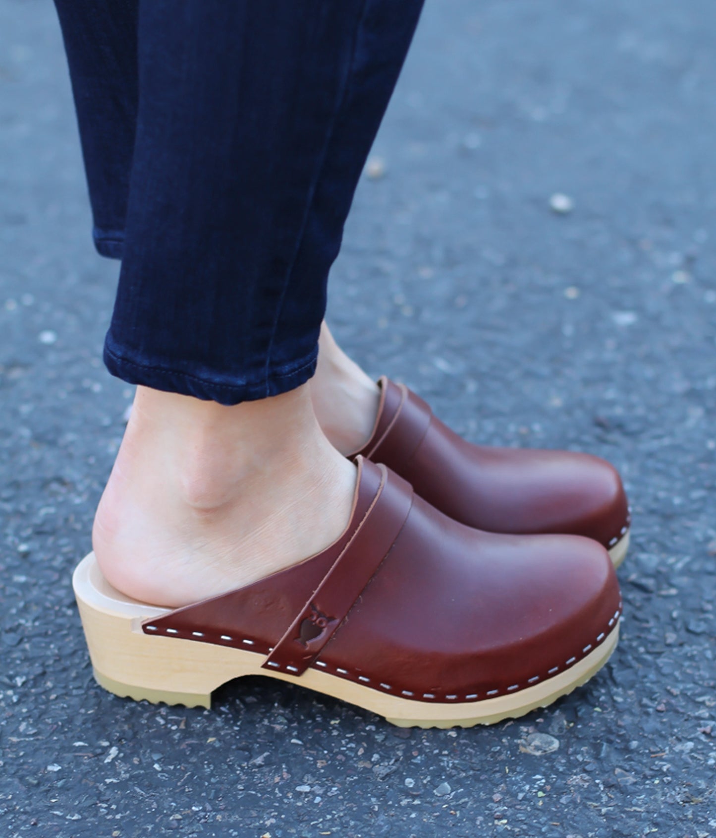 Brown clogs worn on a person's feet with blue jeans on a gray pavement background.classic low heeled clog mule in cognac red vegetable tanned leather stapled on a light wooden base with a leather strap