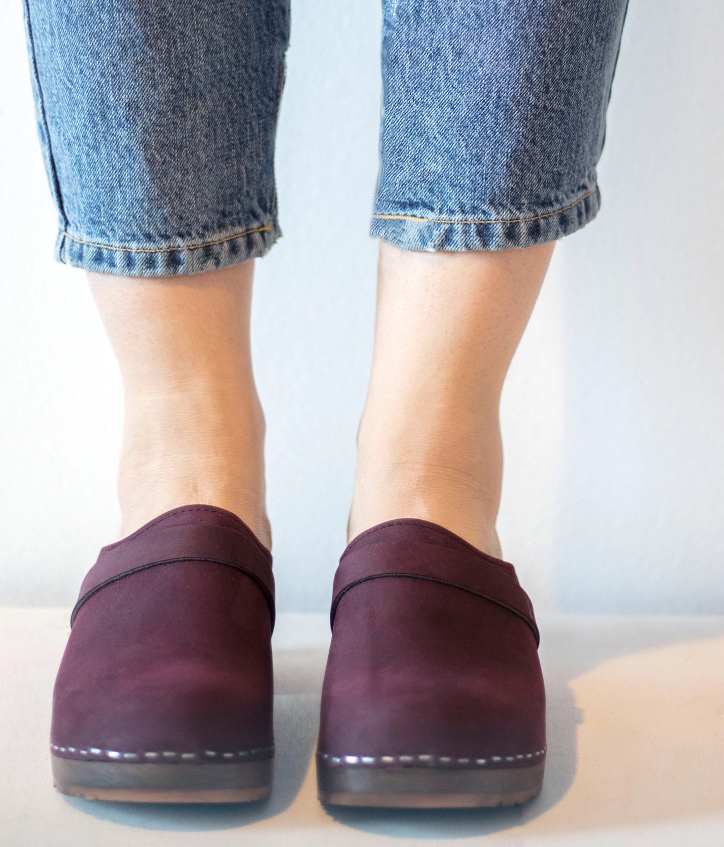Purple clogs worn with blue jeans on a light background.classic low heeled clog mule in plum purple nubuck leather stapled on a dark wooden base with a leather strap