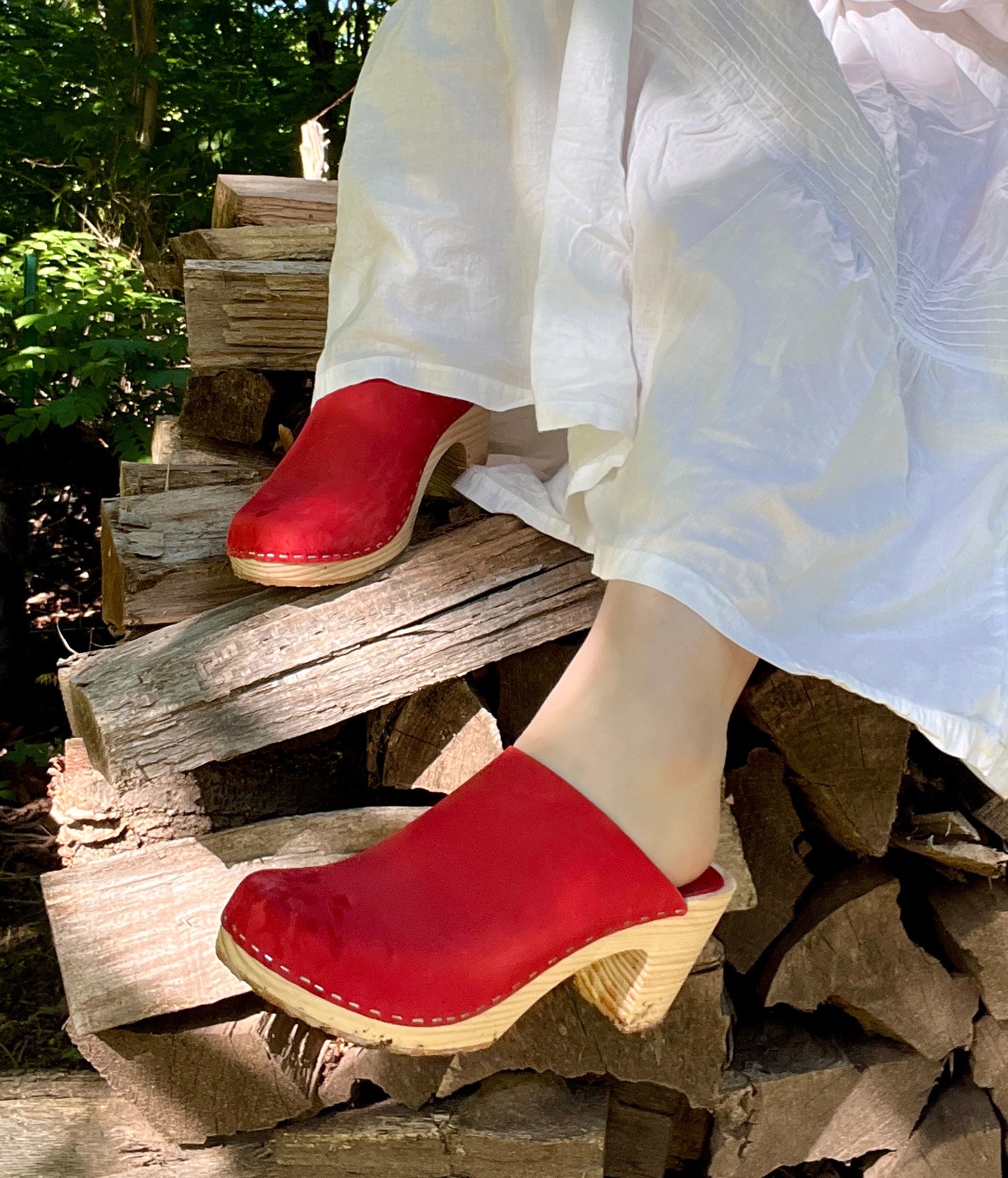 Red clogs worn by a person sitting on wooden logs with a natural background. High rise minimalistic clog mule in red nubuck leather stapled on a light wooden base