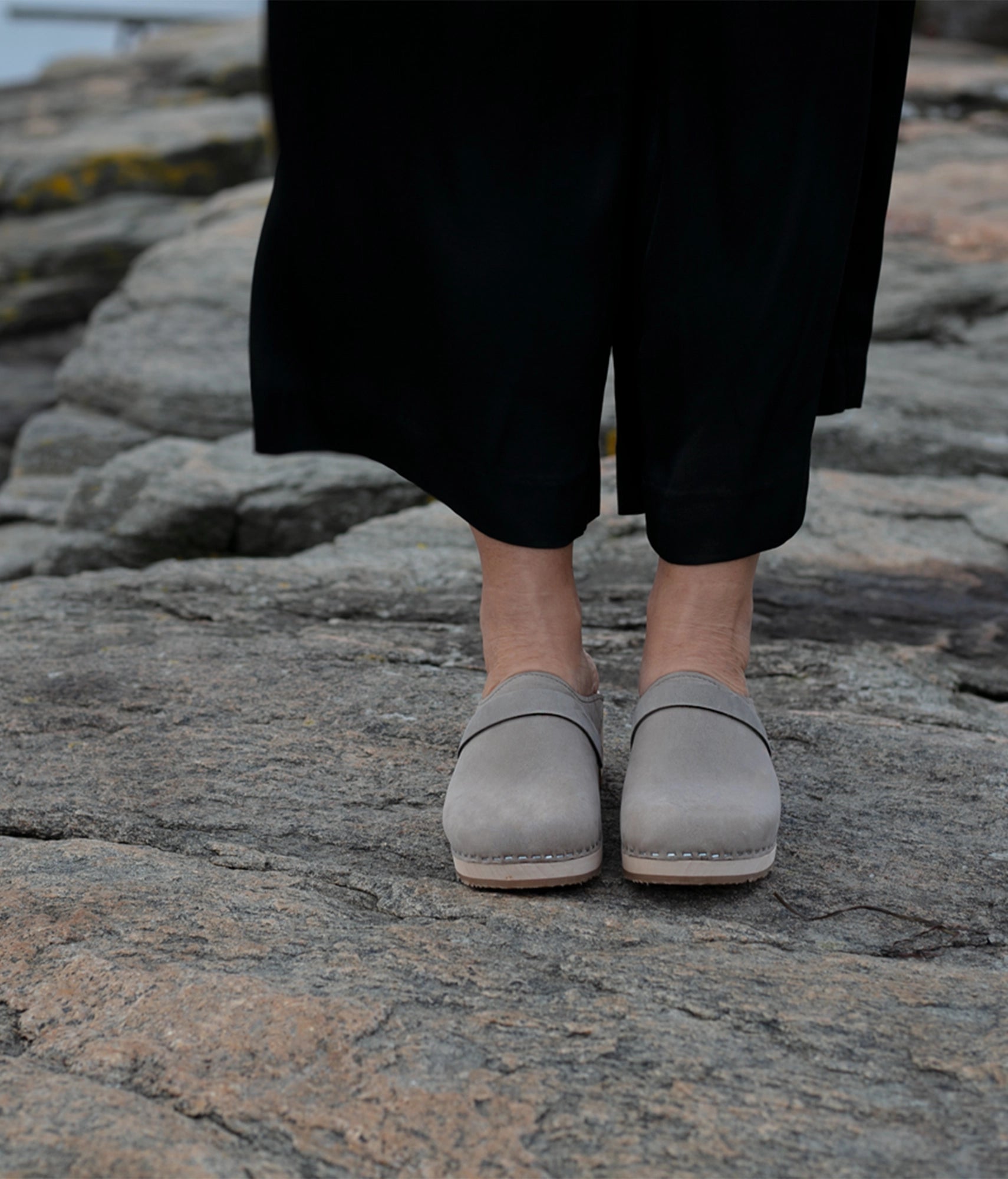 Person wearing gray shoes standing on rocky terrain.classic low heeled clog mule in stone grey nubuck leather stapled on a light wooden base with a leather strap