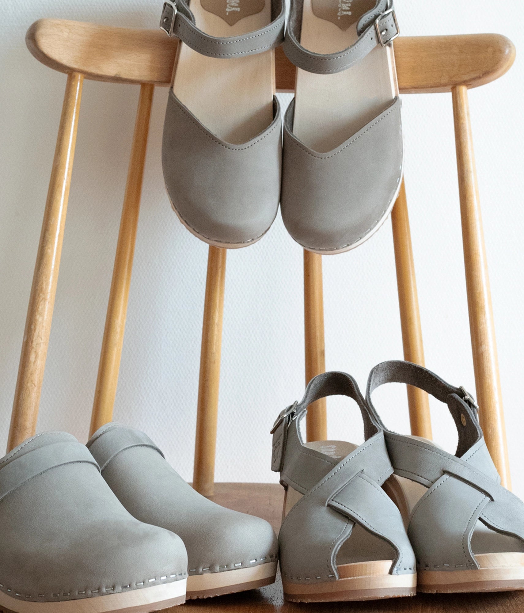 Pairs of gray sandals and clogs on wooden stools against a white wall. classic low heeled clog mule in stone grey nubuck leather stapled on a light wooden base with a leather strap
