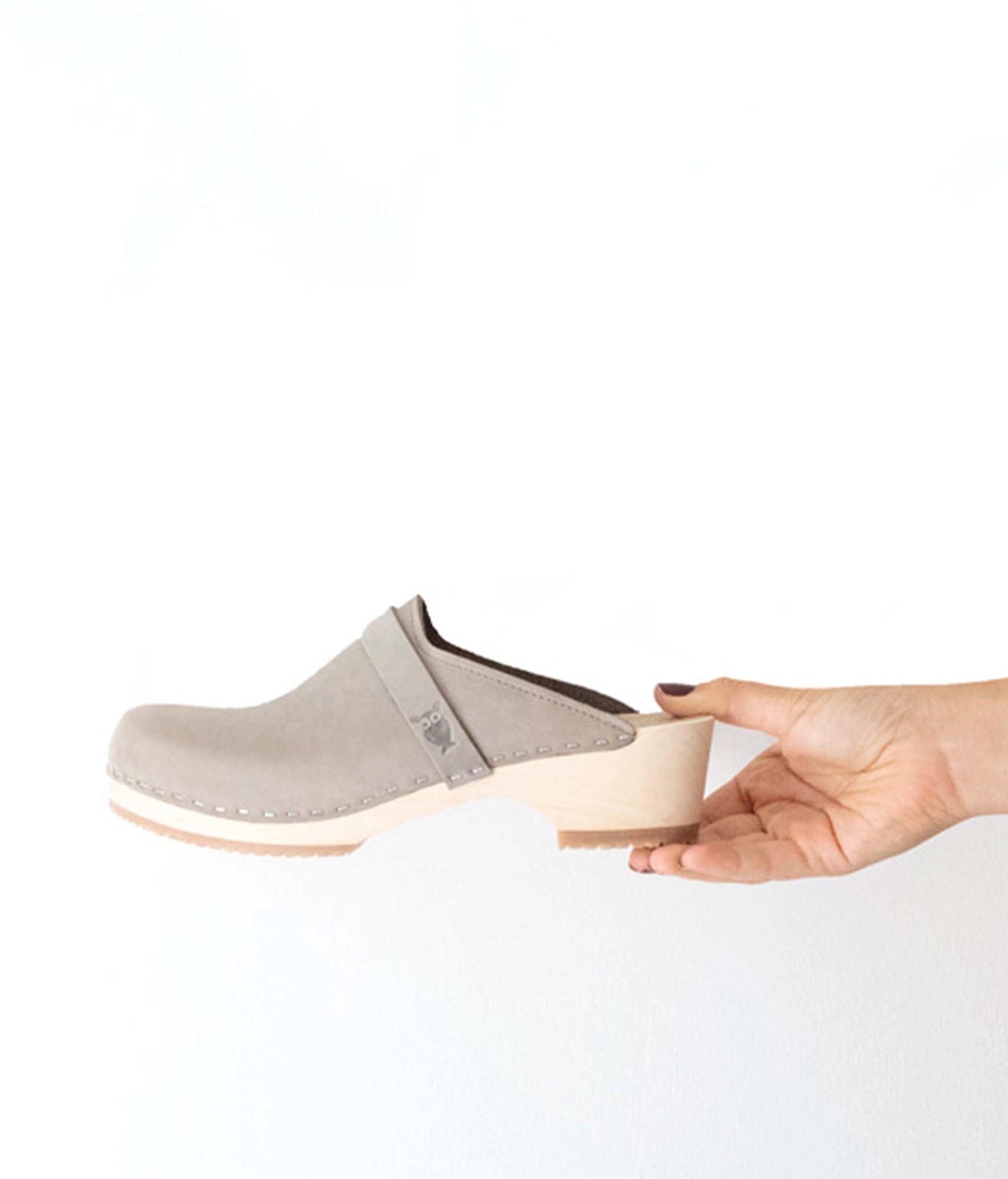 Gray clog shoe with a white sole held by a hand on a white background.classic low heeled clog mule in stone grey nubuck leather stapled on a light wooden base with a leather strap