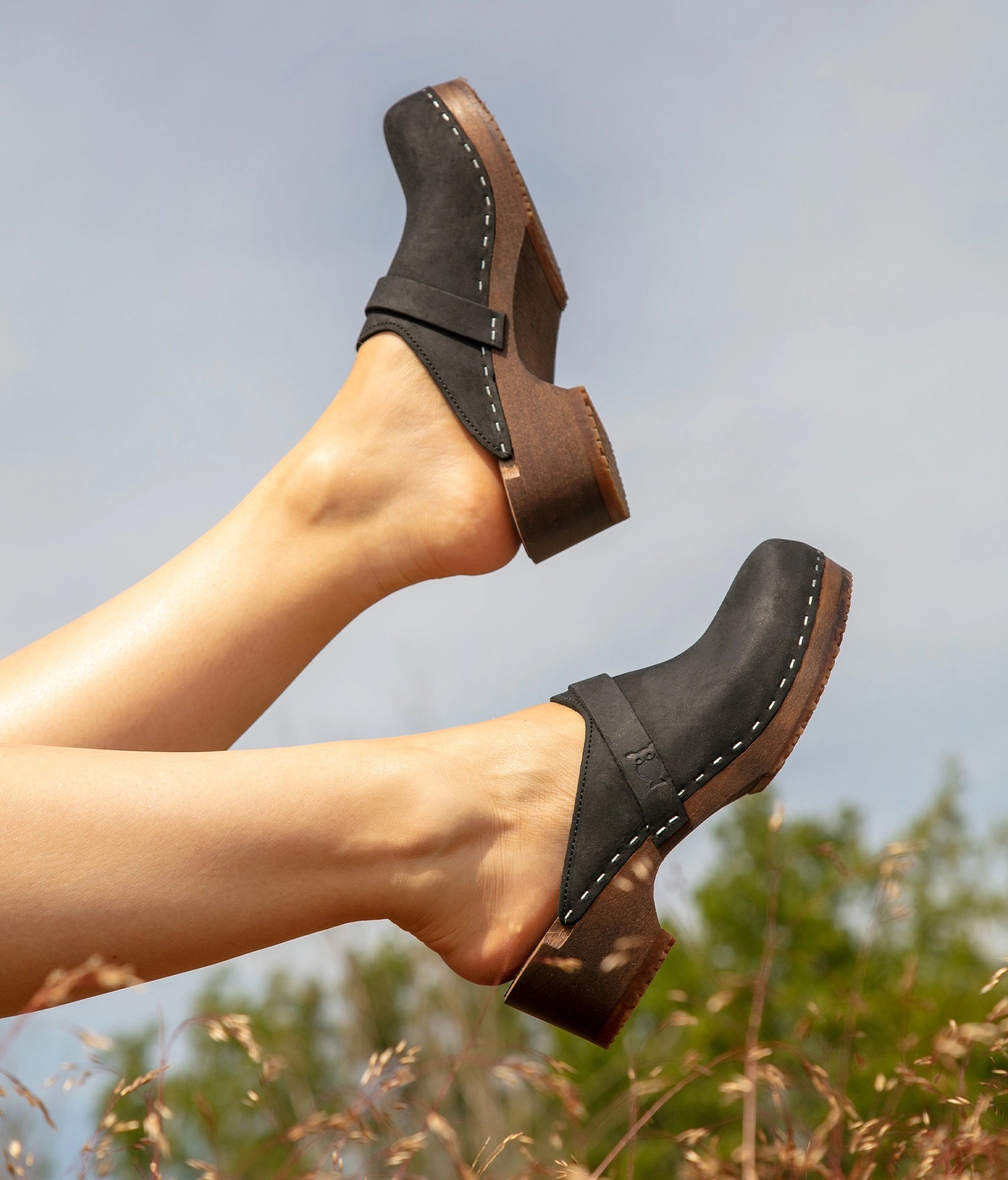 Black clogs worn on feet with a blurred natural background. classic low heeled clog mule in black nubuck leather stapled on a dark wooden base with a leather strap
