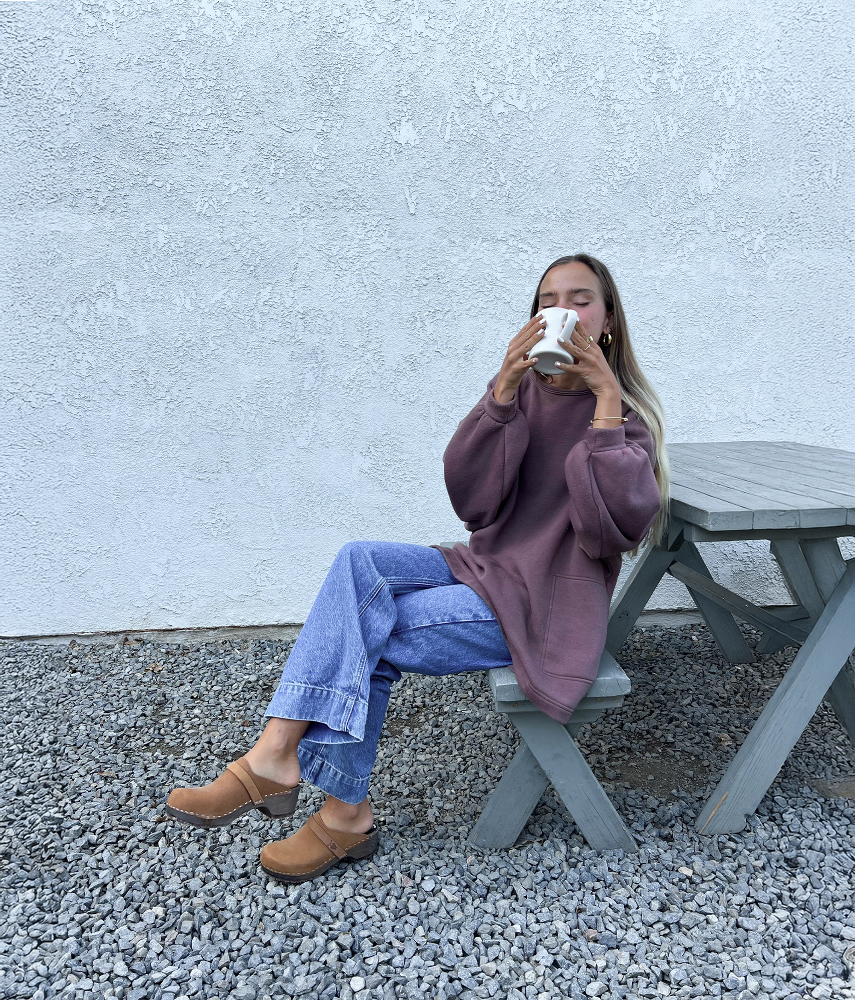 Person sitting on a bench drinking from a cup against a light gray wall. Wearing sandgrens Tokyo clog mules