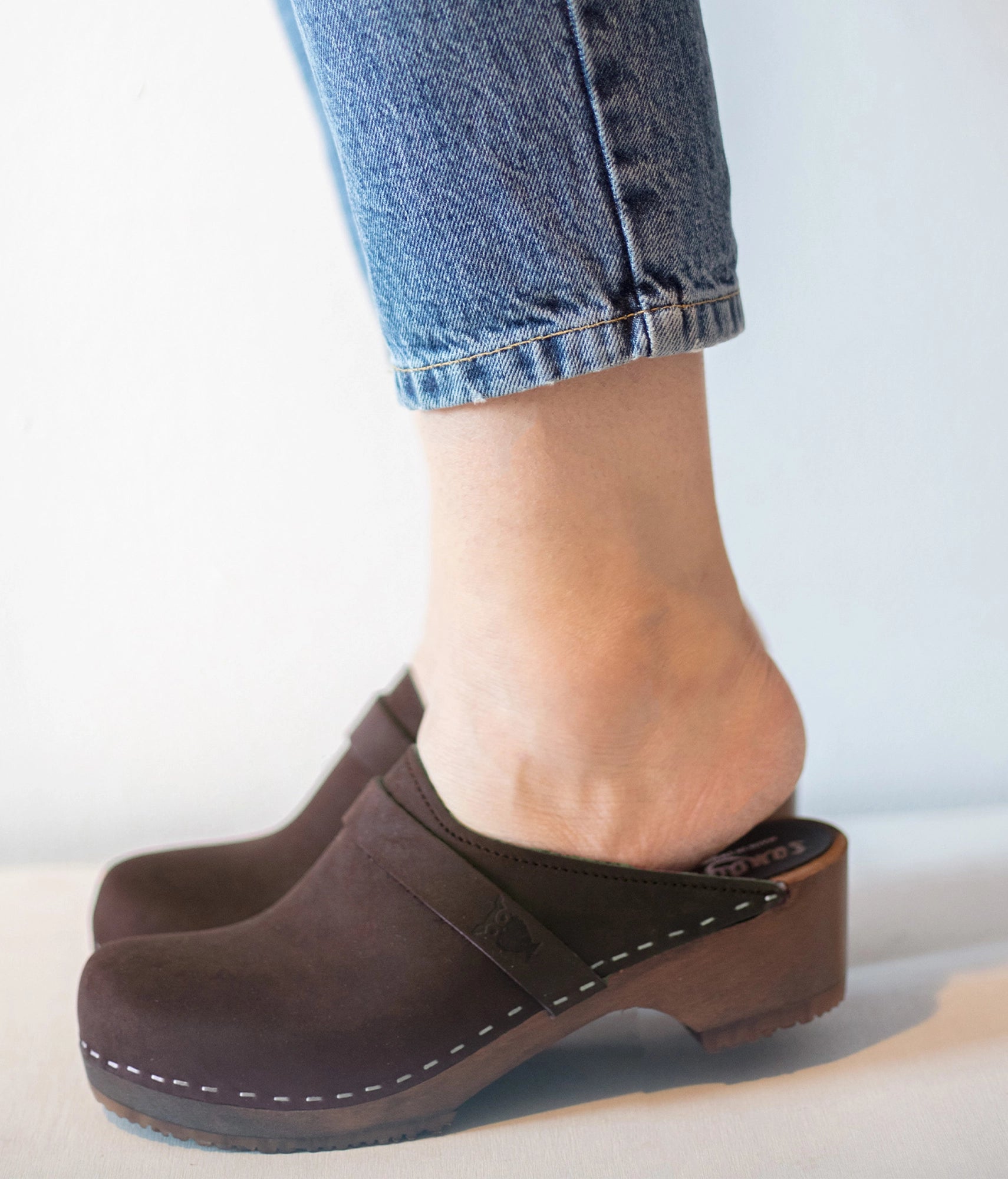 Brown clogs worn with blue jeans on a light background. Classic low heeled clog mule in dark brown nubuck leather stapled on a dark wooden base with a leather strap