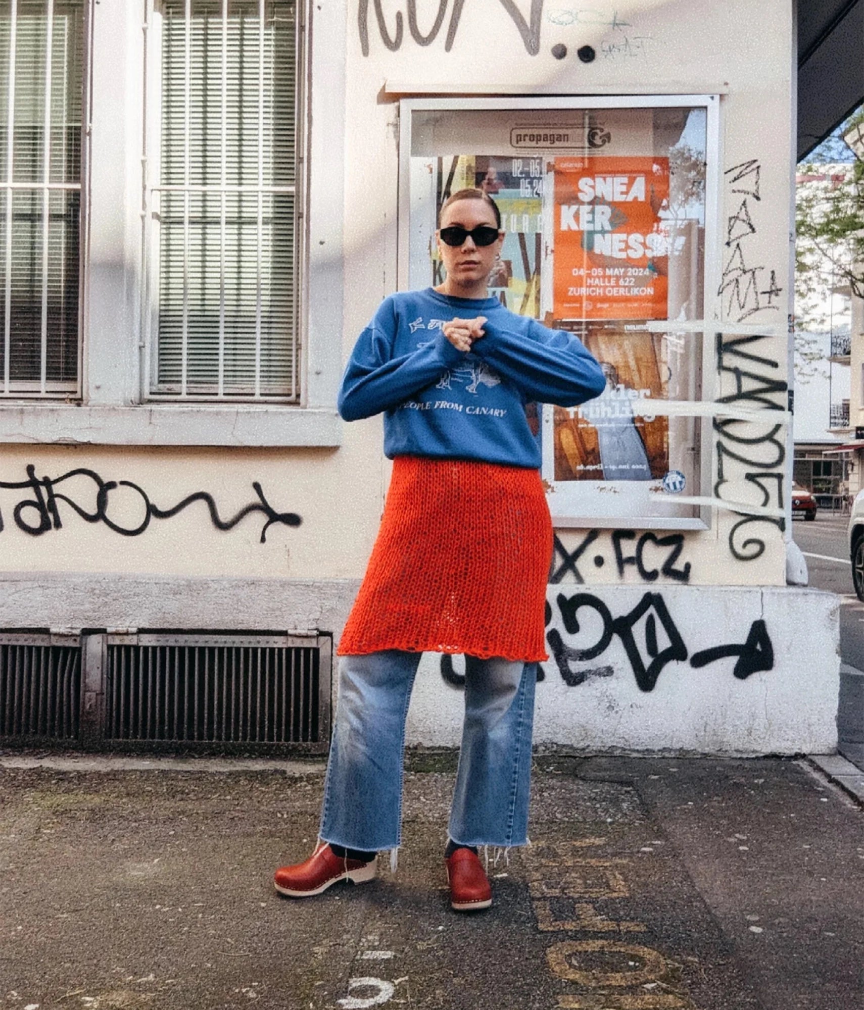Person wearing a blue sweatshirt and red skirt standing in front of a graffiti-covered wall. classic low heeled clog mule in red vegetable tanned leather stapled on a light wooden base with a leather strap