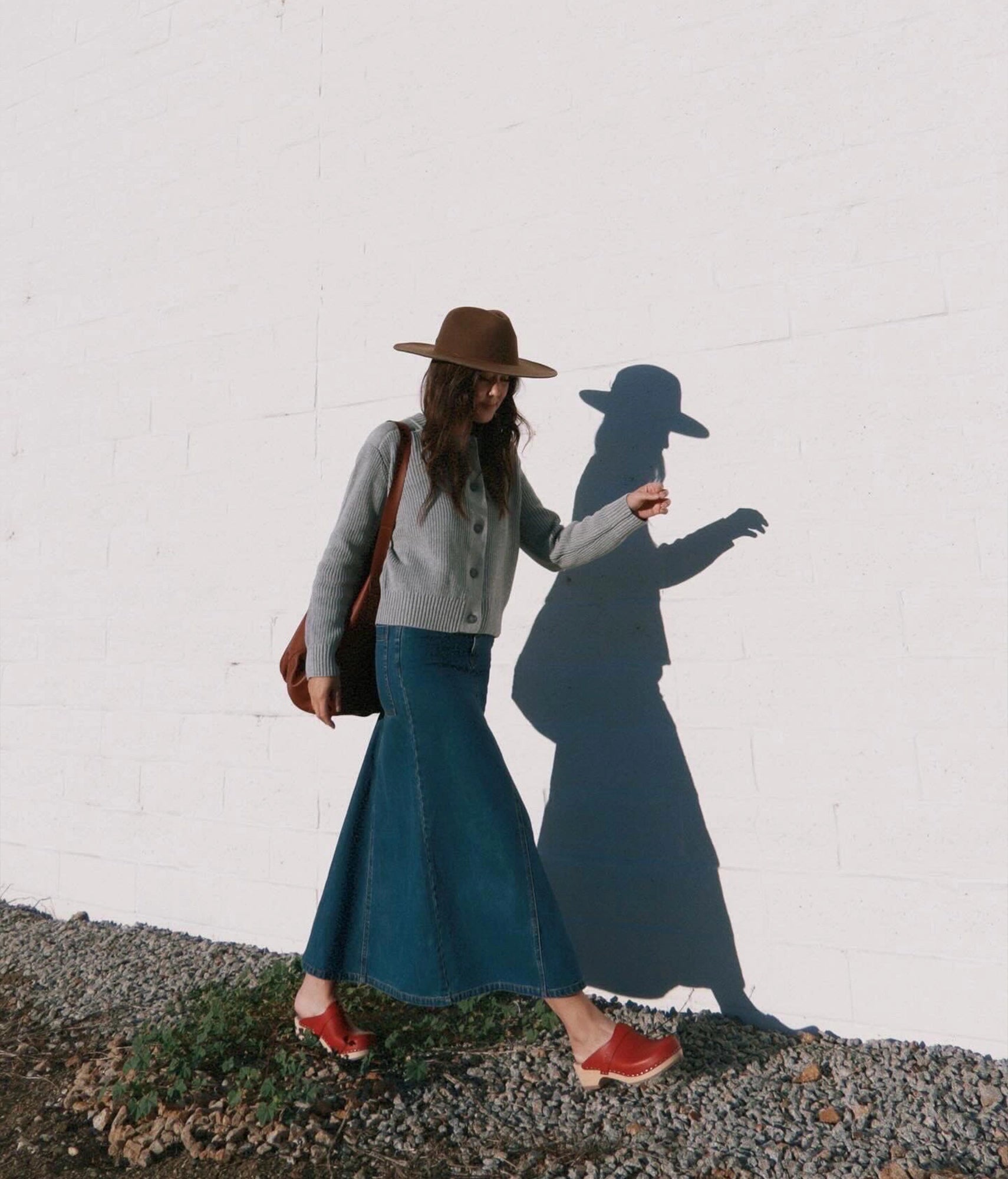 Woman in a blue skirt and brown hat standing against a white brick wall with her shadow visible. classic low heeled clog mule in red vegetable tanned leather stapled on a light wooden base with a leather strap