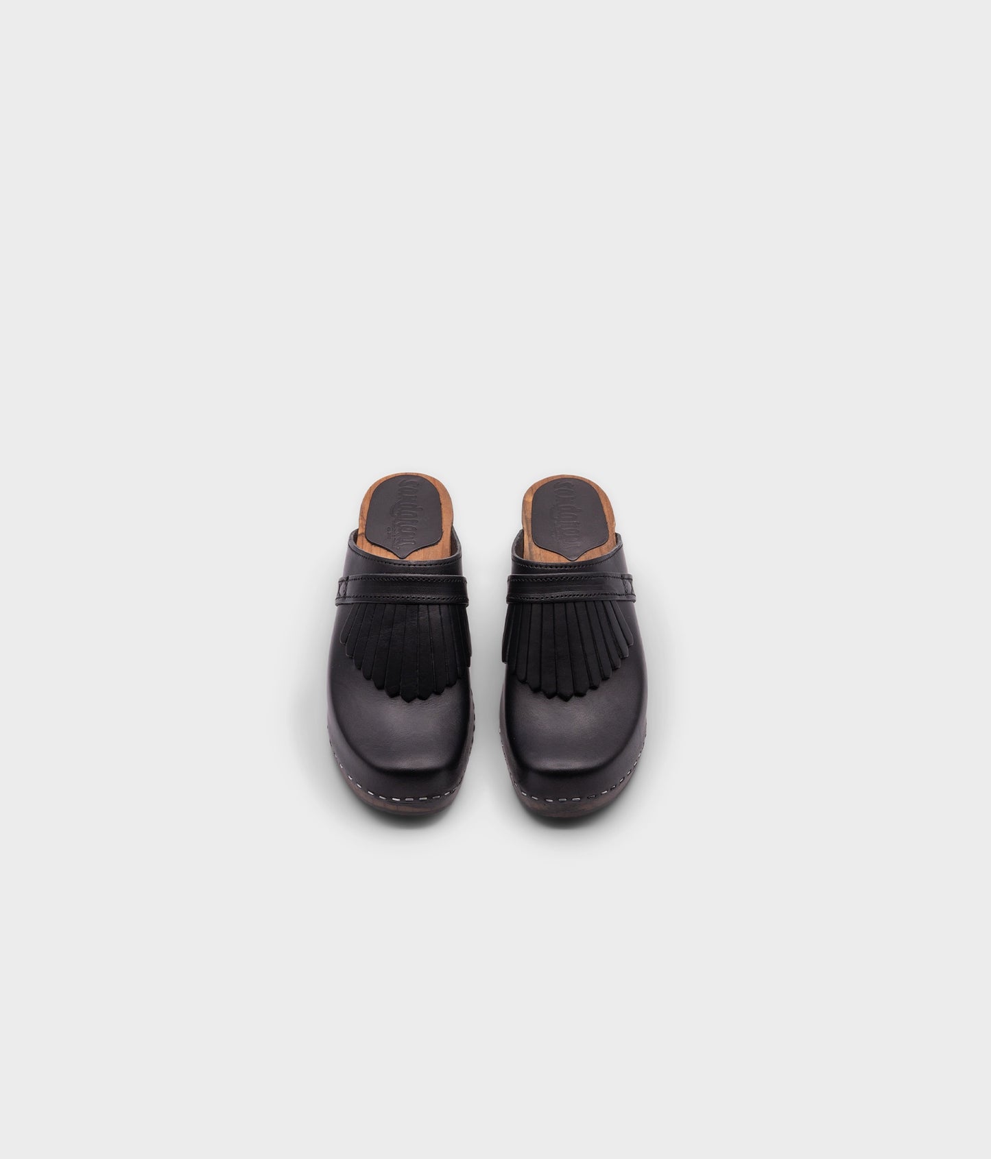high rise clog mule in black vegetable tanned leather stapled on a dark wooden base with a leather strap with fringes