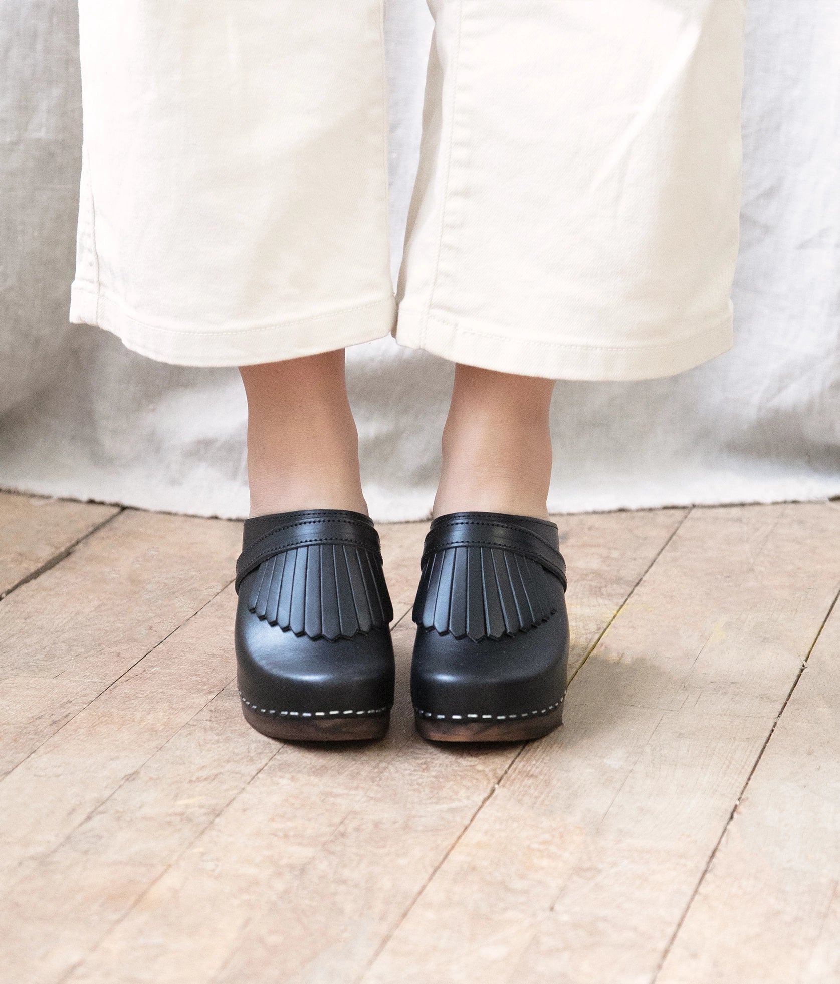 high rise clog mule in black vegetable tanned leather stapled on a dark wooden base with a leather strap with fringes