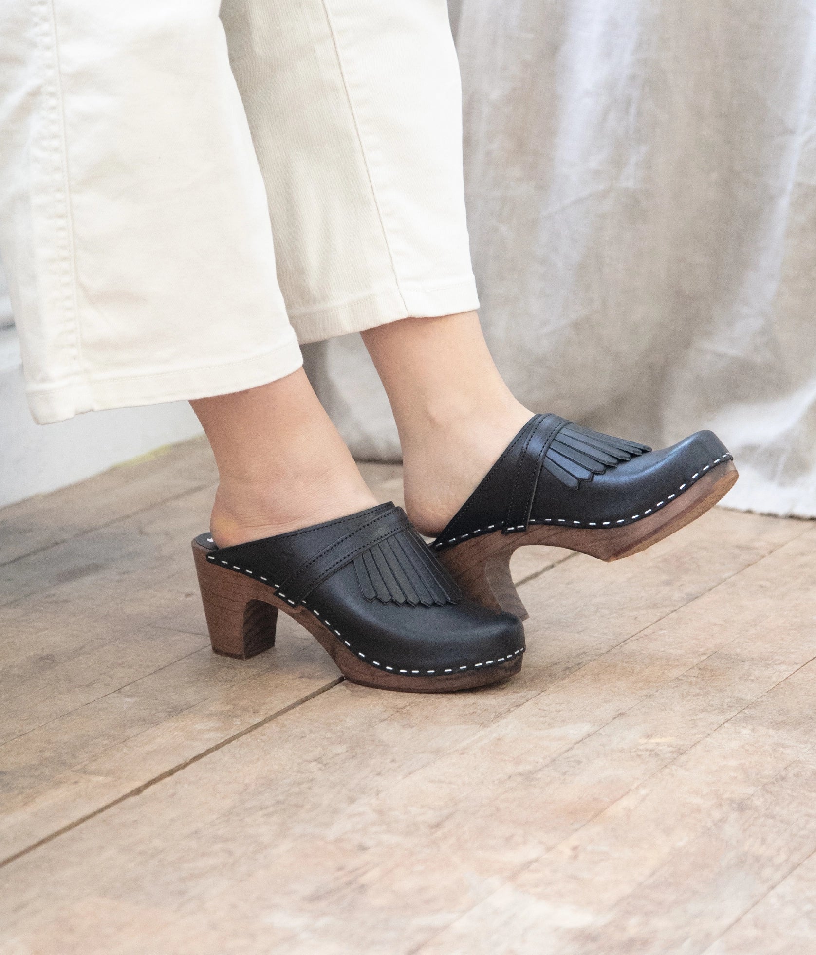 high rise clog mule in black vegetable tanned leather stapled on a dark wooden base with a leather strap with fringes