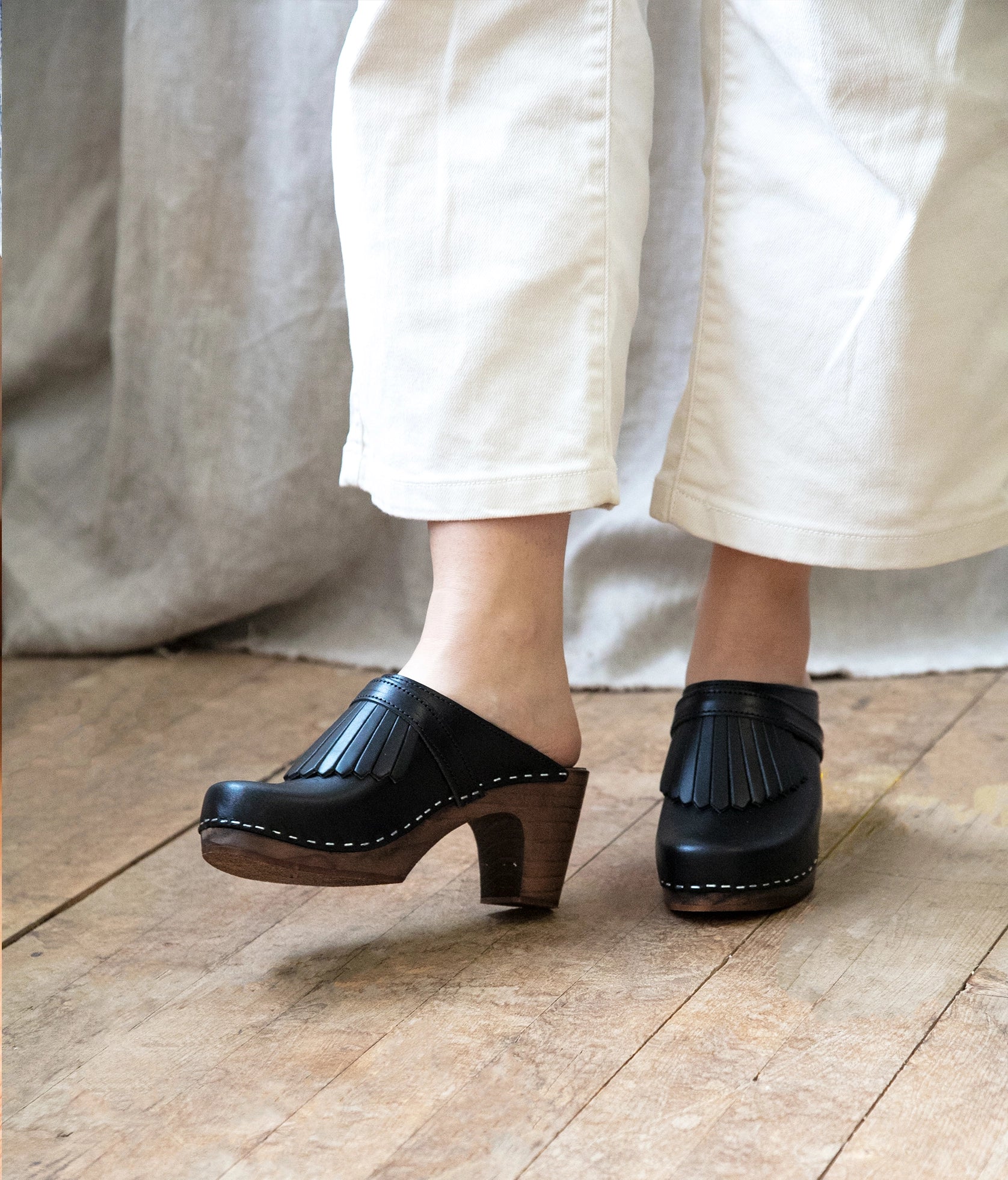 high rise clog mule in black vegetable tanned leather stapled on a dark wooden base with a leather strap with fringes