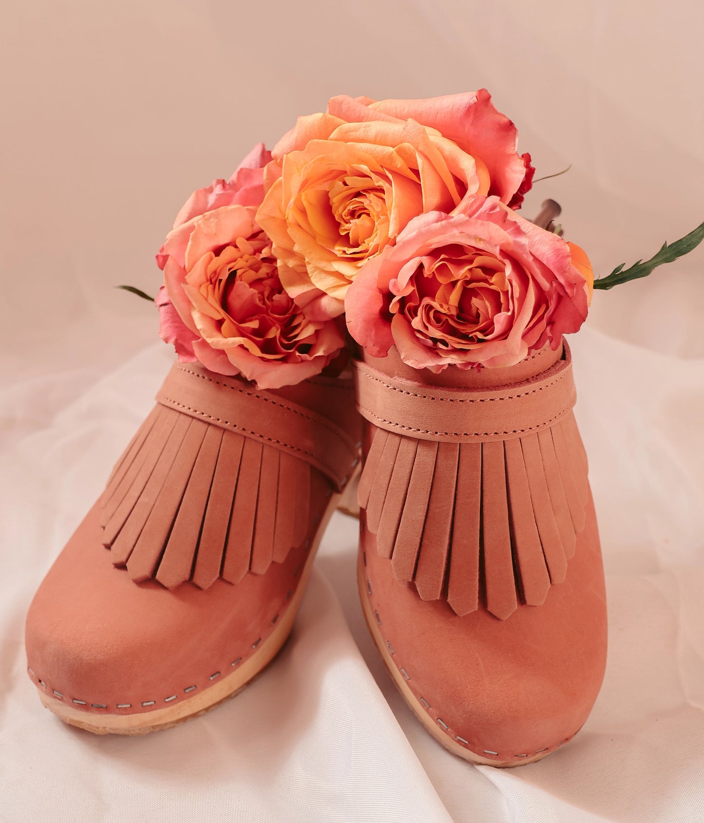 Pair of pink fringed shoes with flowers on a light background. High rise clog mule in blush pink nubuck leather stapled on a light wooden base with a leather strap with fringes