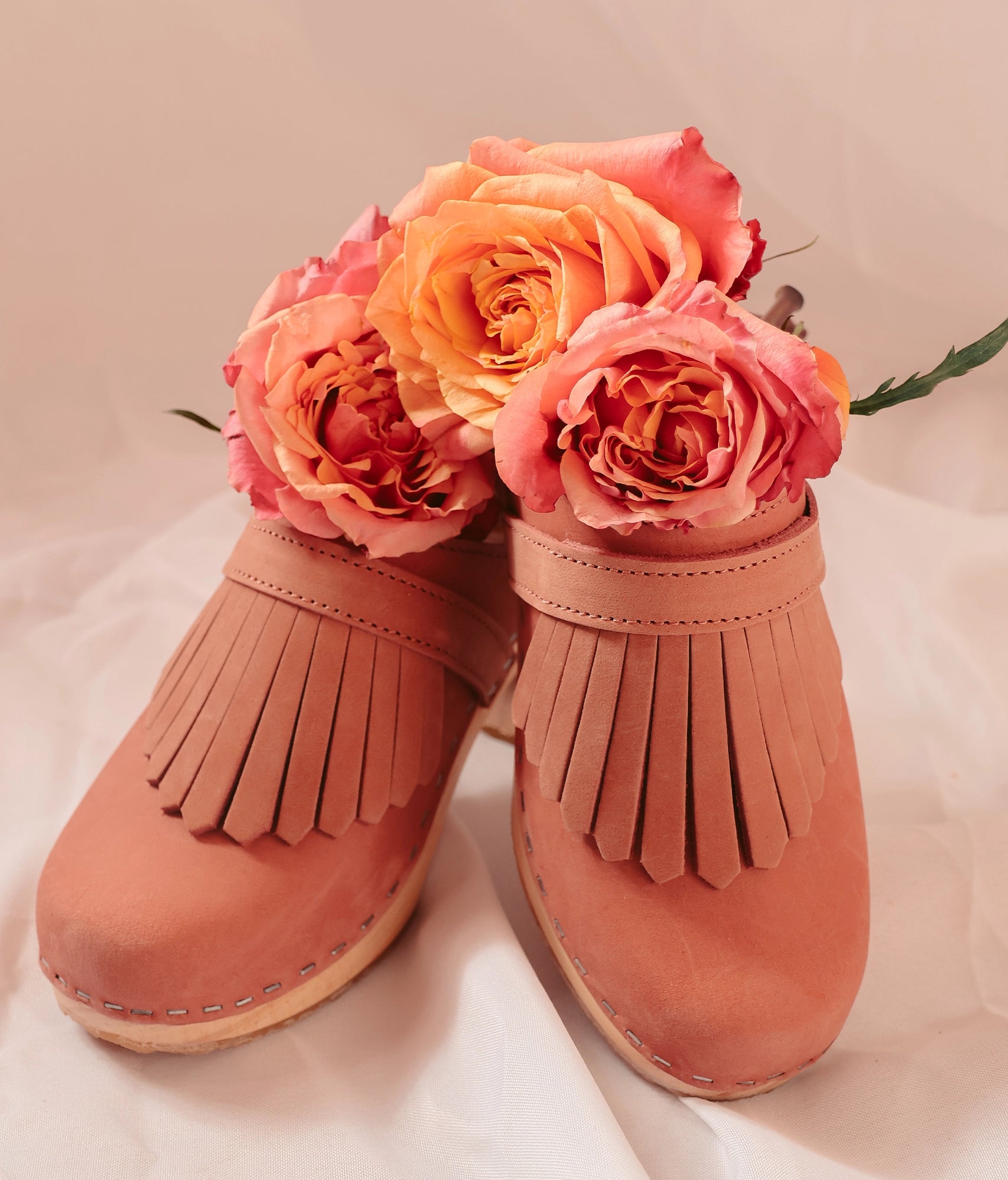 Pair of pink fringed shoes with flowers on a light background. High rise clog mule in blush pink nubuck leather stapled on a light wooden base with a leather strap with fringes