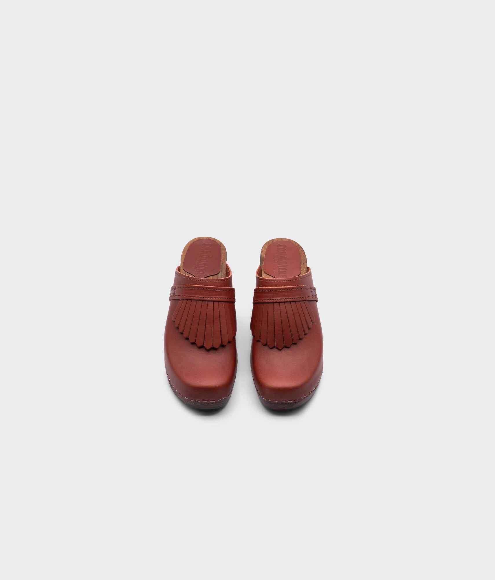 high rise clog mule in cognac red vegetable tanned leather stapled on a dark wooden base with a leather strap with fringes