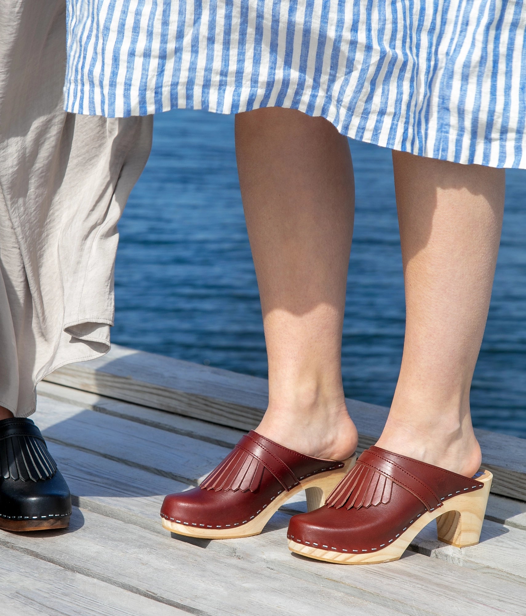Person wearing cognac colored clogs with wooden soles on a dock by water. high rise clog mule in cognac red vegetable tanned leather stapled on a light wooden base with a leather strap with fringes