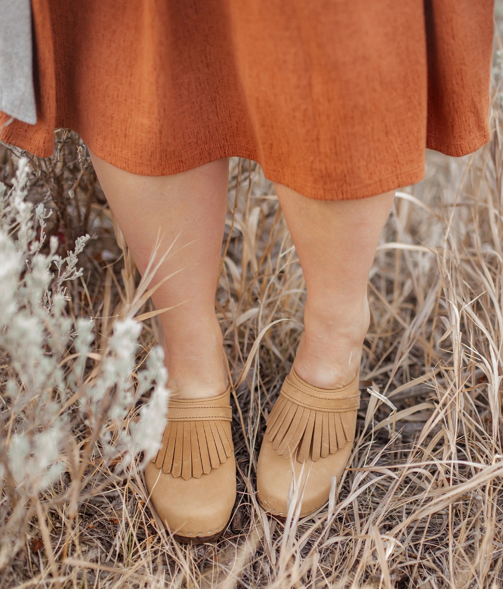 high rise clog mule in light brown nubuck leather stapled on a dark wooden base with a leather strap with fringes
