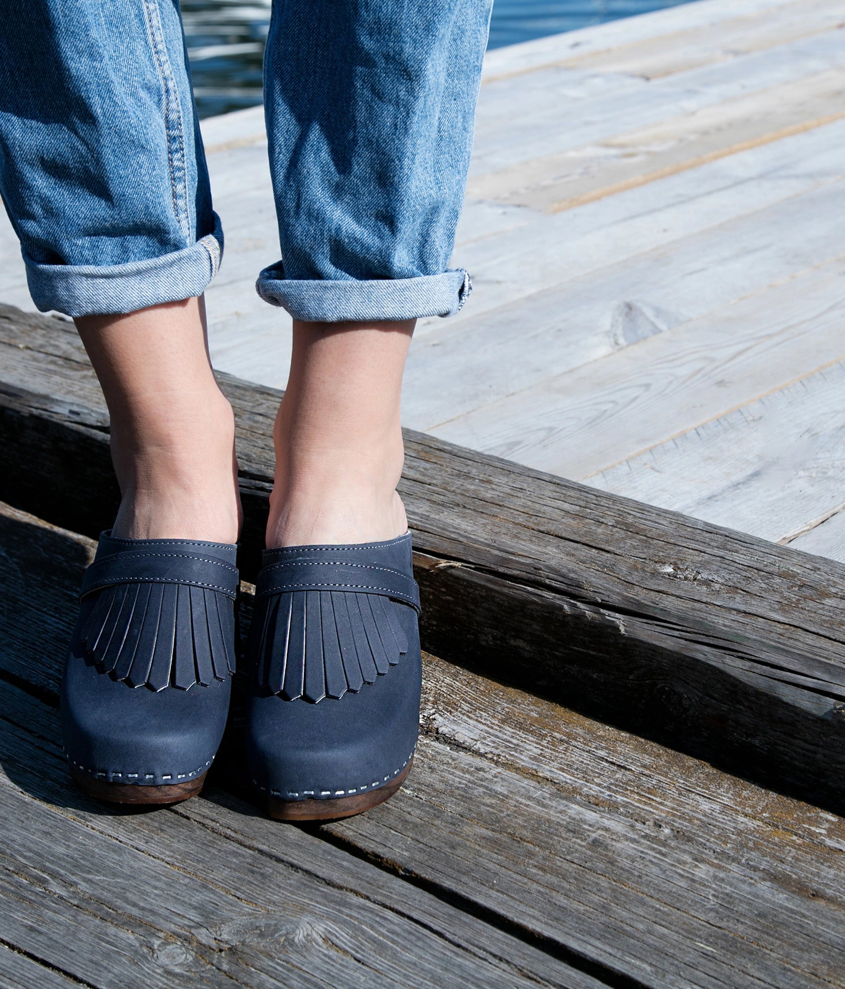 Navy blue clogs with fringes worn by a person on a wooden dock. High rise clog mule in navy blue nubuck leather stapled on a dark wooden base with a leather strap with fringes