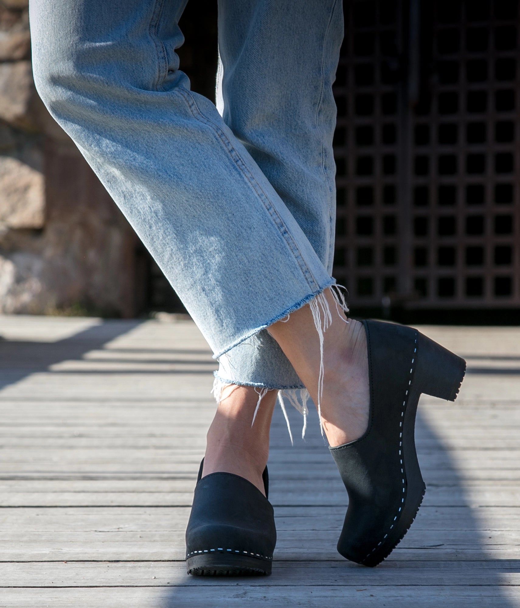 Person wearing black clogs and light blue jeans on a wooden floor. High heeled closed-back clogs in black nubuck leather stapled on a black wooden base