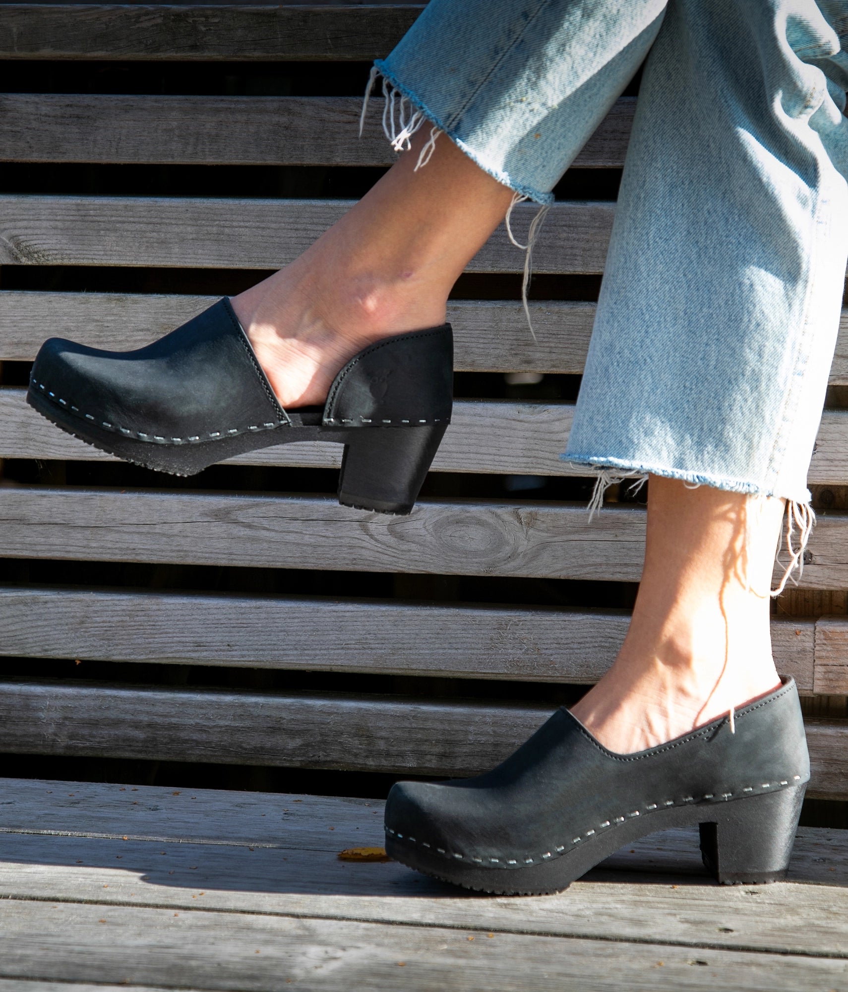 Person wearing black clogs on a wooden floor. High heeled closed-back clogs in black nubuck leather stapled on a dark wooden base