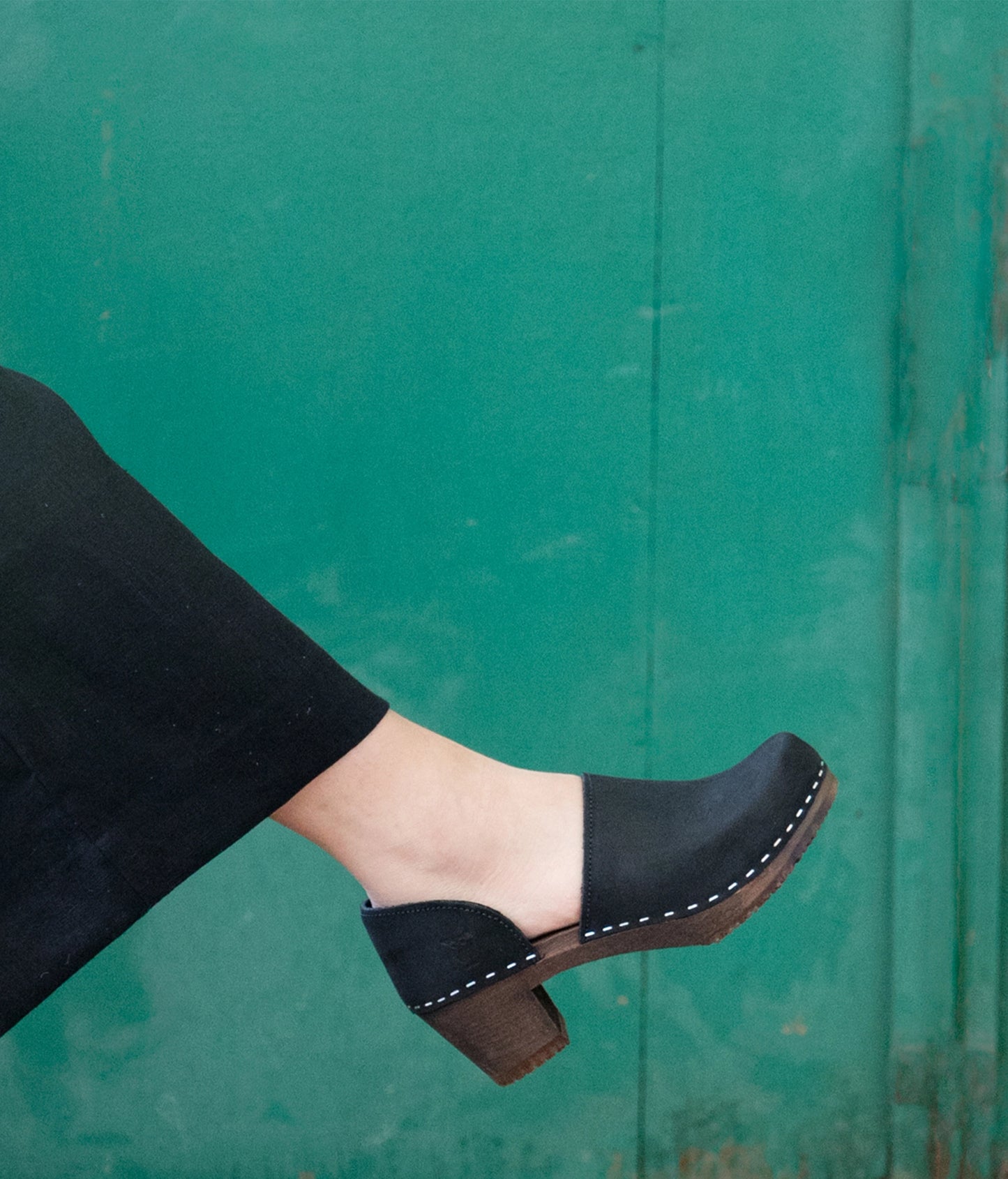 Black clog shoe on a foot against a green background. high heeled closed-back clogs in black nubuck leather stapled on a dark wooden base