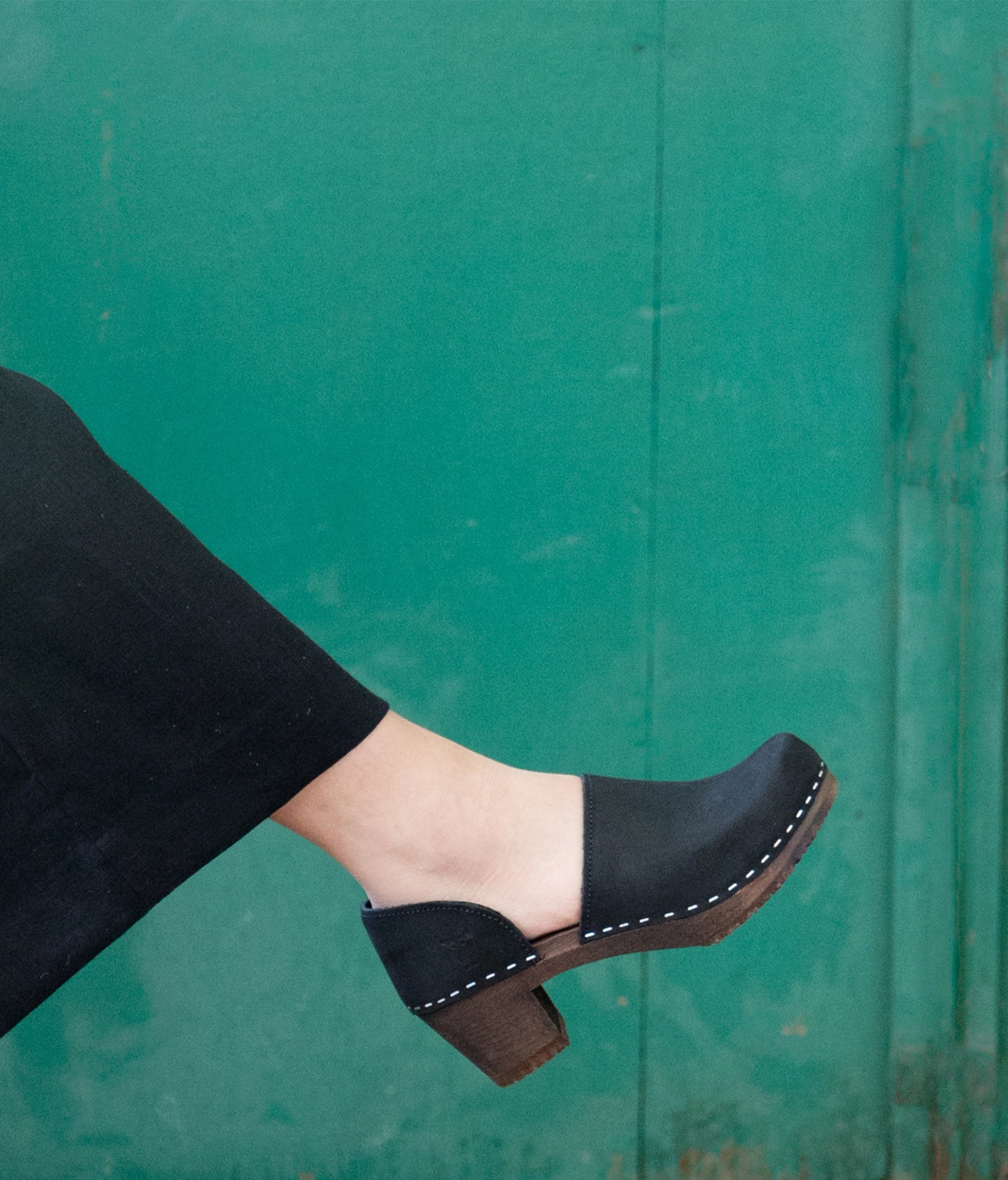 Black clog shoe on a foot against a green background. high heeled closed-back clogs in black nubuck leather stapled on a dark wooden base