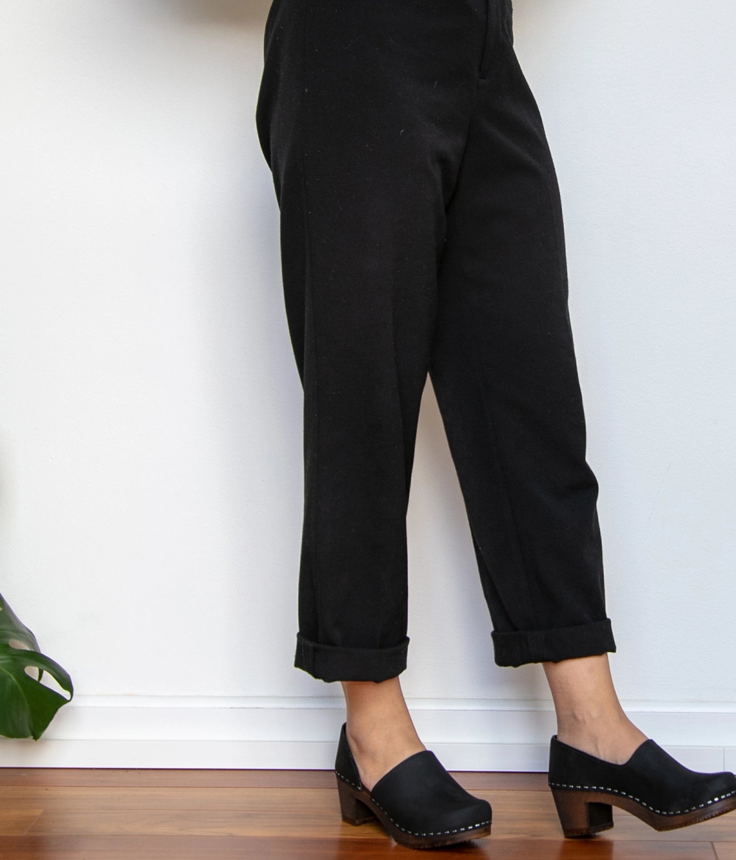 Person wearing black pants and clogs against a white wall. High heeled closed-back clogs in black nubuck leather stapled on a dark wooden base