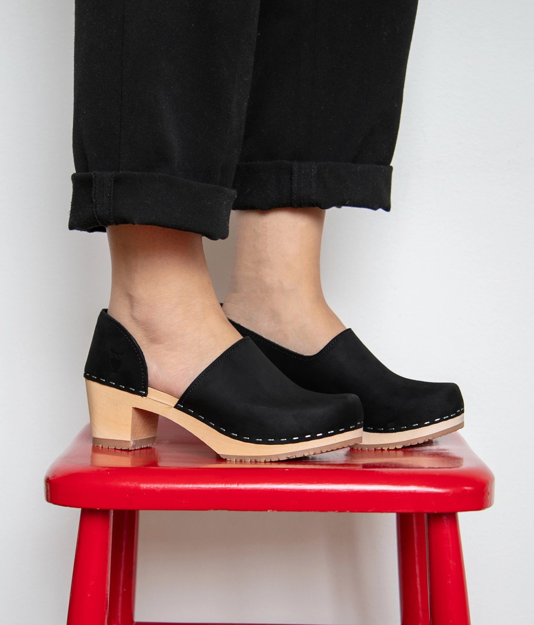 Black closed-back clogs worn by a person on a red stool against a white background. High heeled closed-back clogs in black nubuck leather stapled on a light wooden base