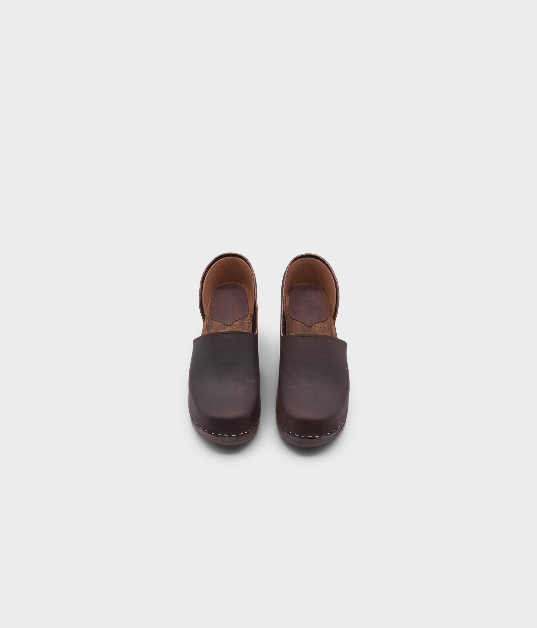 high heeled closed-back clogs in dark brown nubuck leather stapled on a dark wooden base