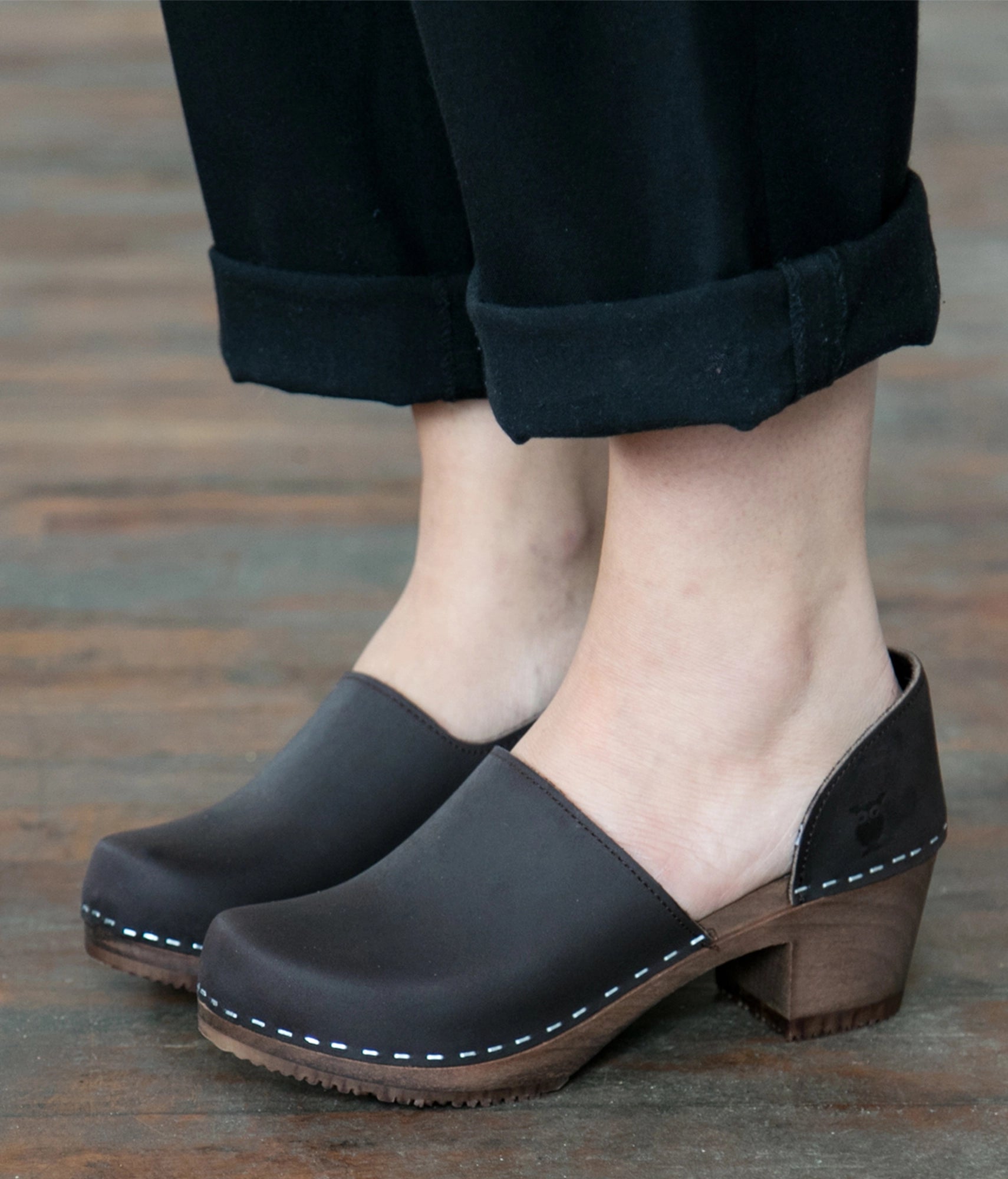 Fudge clogs with wooden soles worn by a person on a wooden floor. High heeled closed-back clogs in dark brown nubuck leather stapled on a dark wooden base