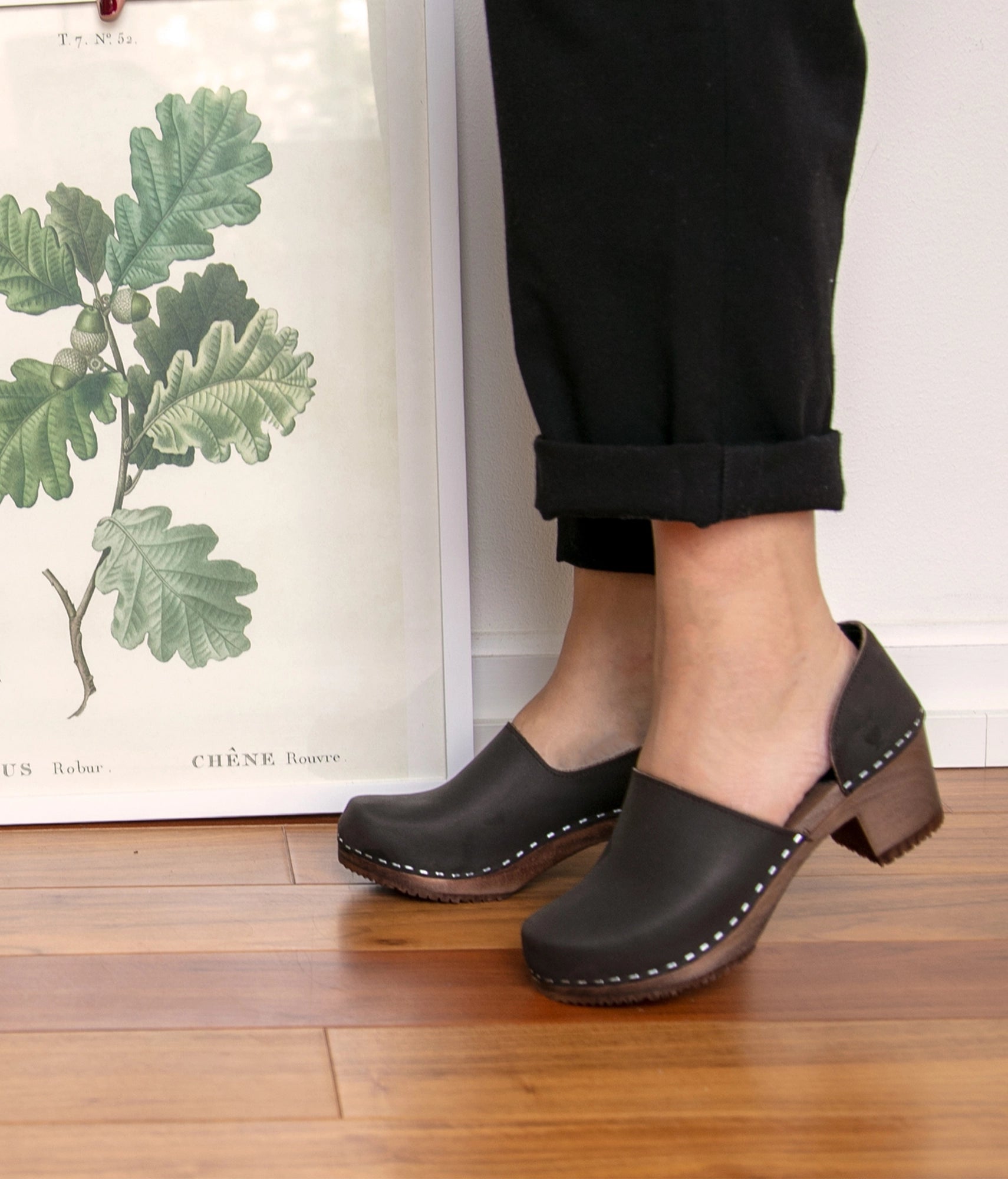 Person wearing black clogs on a wooden floor with a plant poster in the background. high heeled closed-back clogs in dark brown nubuck leather stapled on a dark wooden base