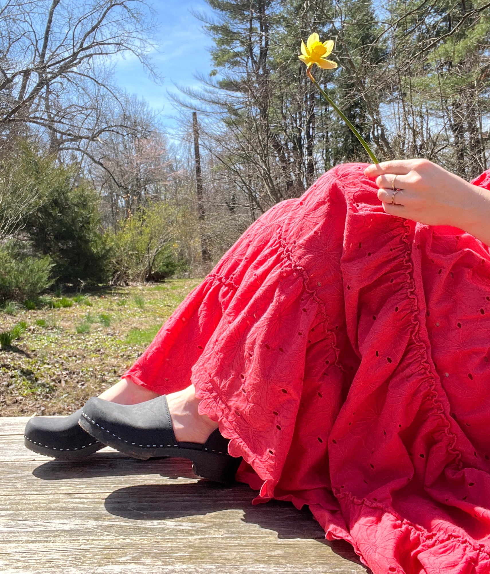 Person in a red dress and black shoes sitting on a wooden deck holding a yellow flower. Low heeled closed-back clogs in black nubuck leather stapled on a black wooden base