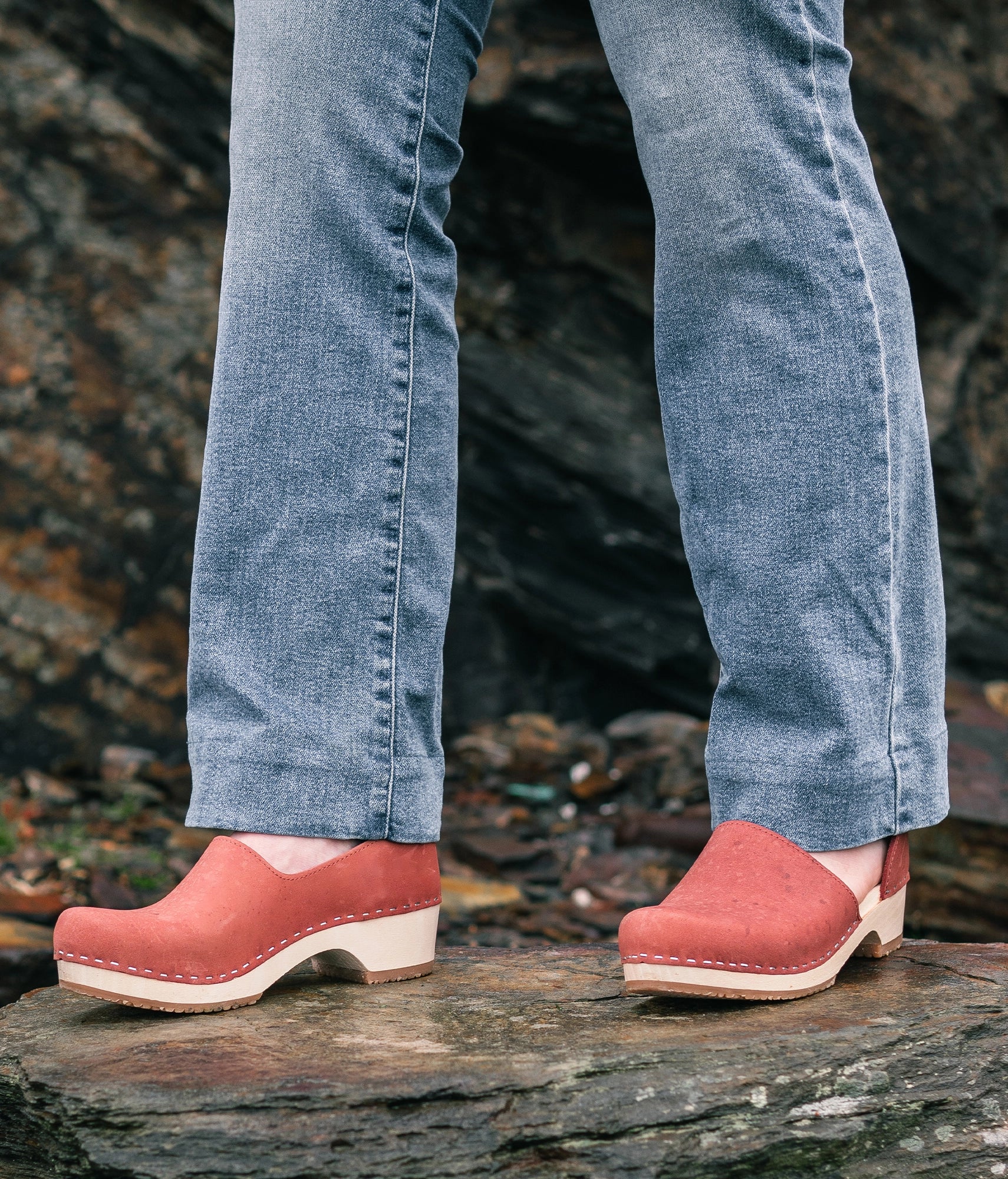 Person wearing red clogs and blue jeans standing on a rocky surface. low heeled closed-back clogs in blush pink nubuck leather stapled on a light wooden base