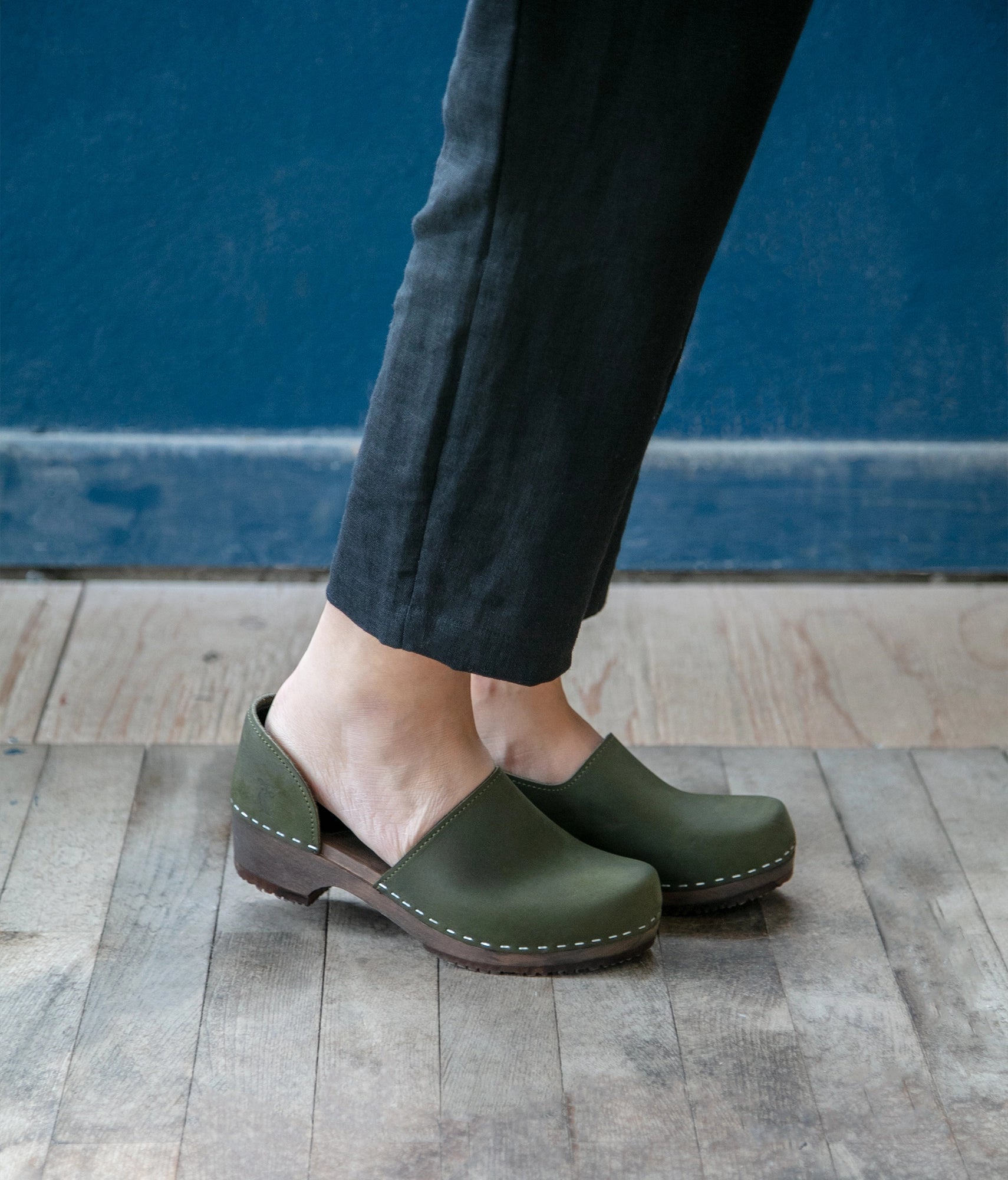 low heeled closed-back clogs in olive green nubuck leather stapled on a dark wooden base