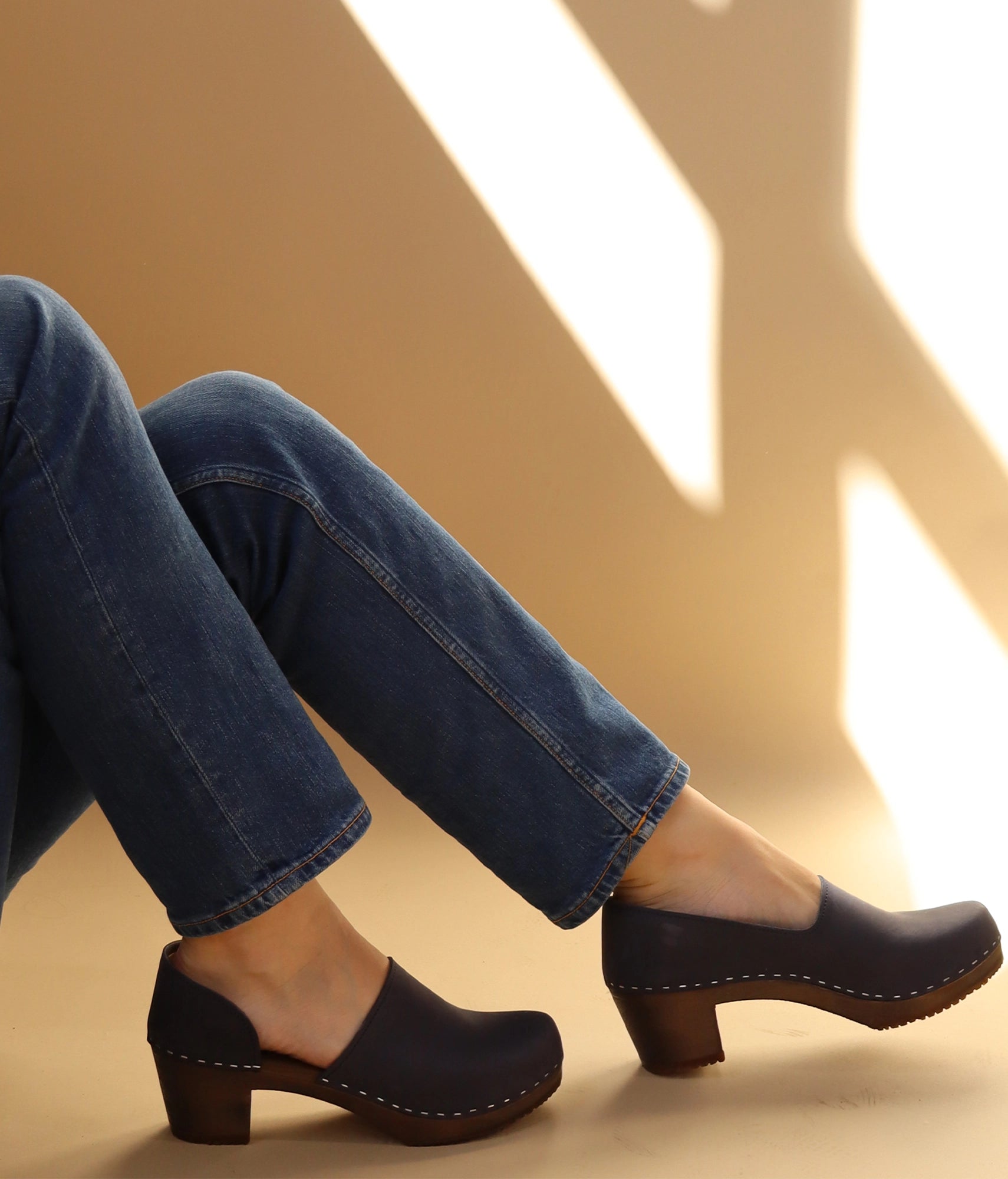 Person wearing Navy colored clogs and blue jeans on a beige background. high heeled closed-back clogs in navy blue nubuck leather stapled on a dark wooden base
