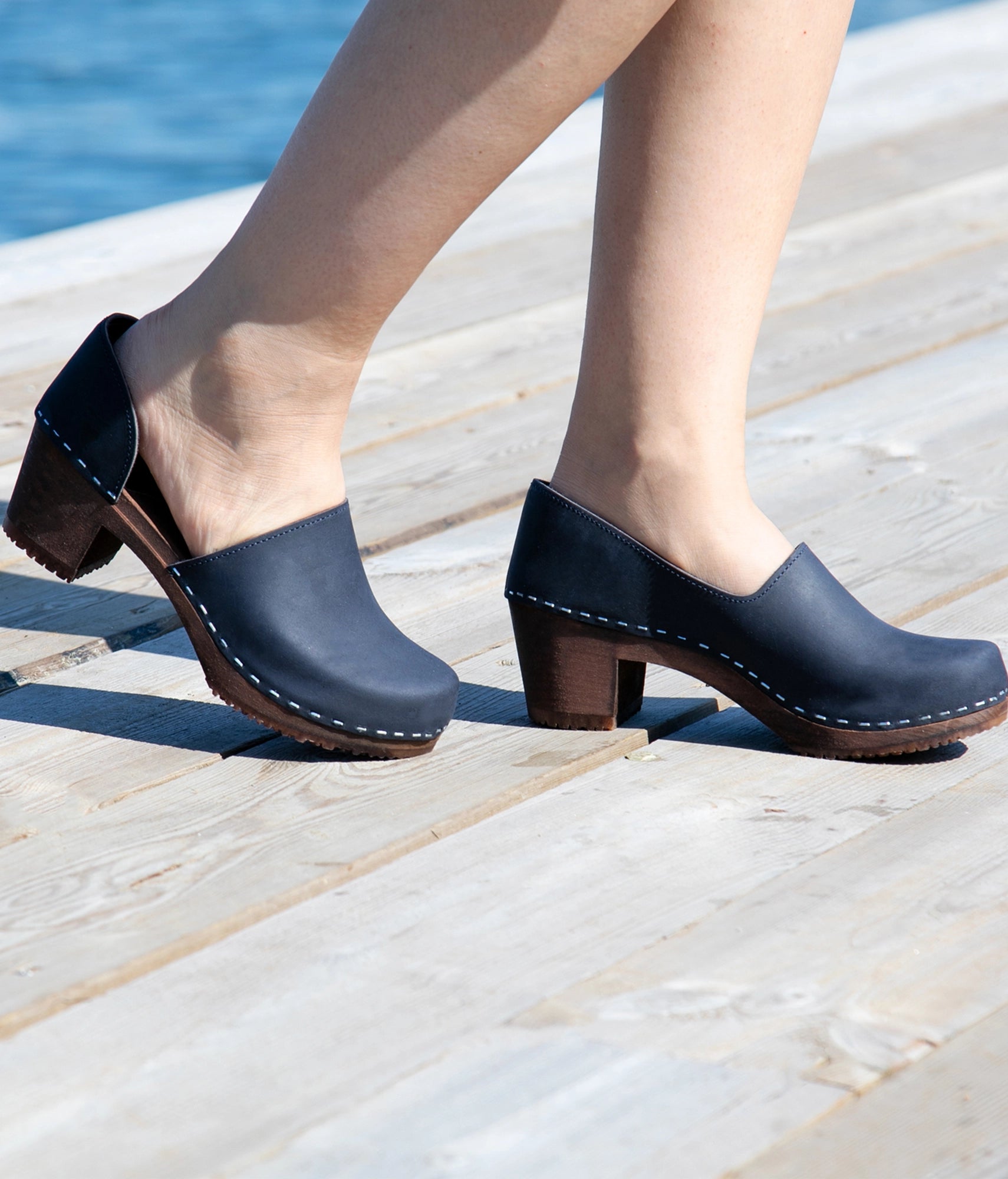 Navy blue clogs with wooden soles worn by a person on a wooden deck. High heeled closed-back clogs in navy blue nubuck leather stapled on a dark wooden base