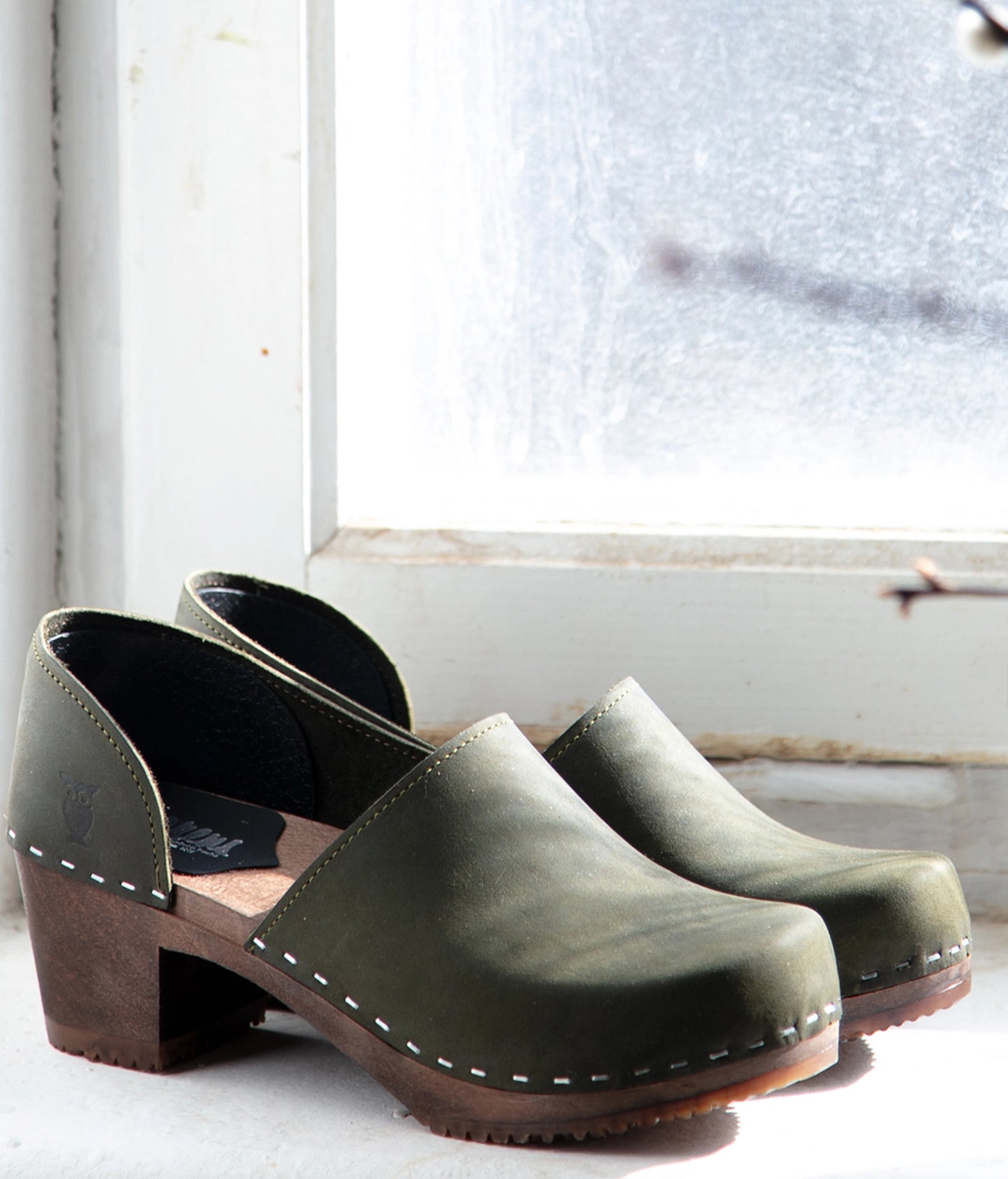 Pair of green wooden clogs on a white surface with a window in the background. High heeled closed-back clogs in olive green nubuck leather stapled on a dark wooden base