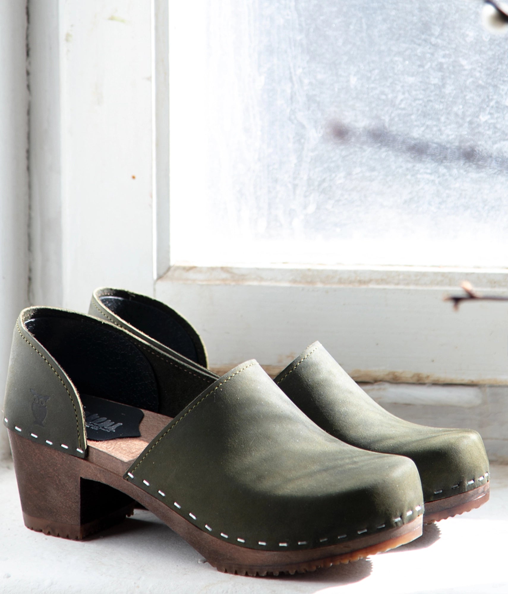 Pair of green wooden clogs on a white surface with a window in the background. High heeled closed-back clogs in olive green nubuck leather stapled on a dark wooden base