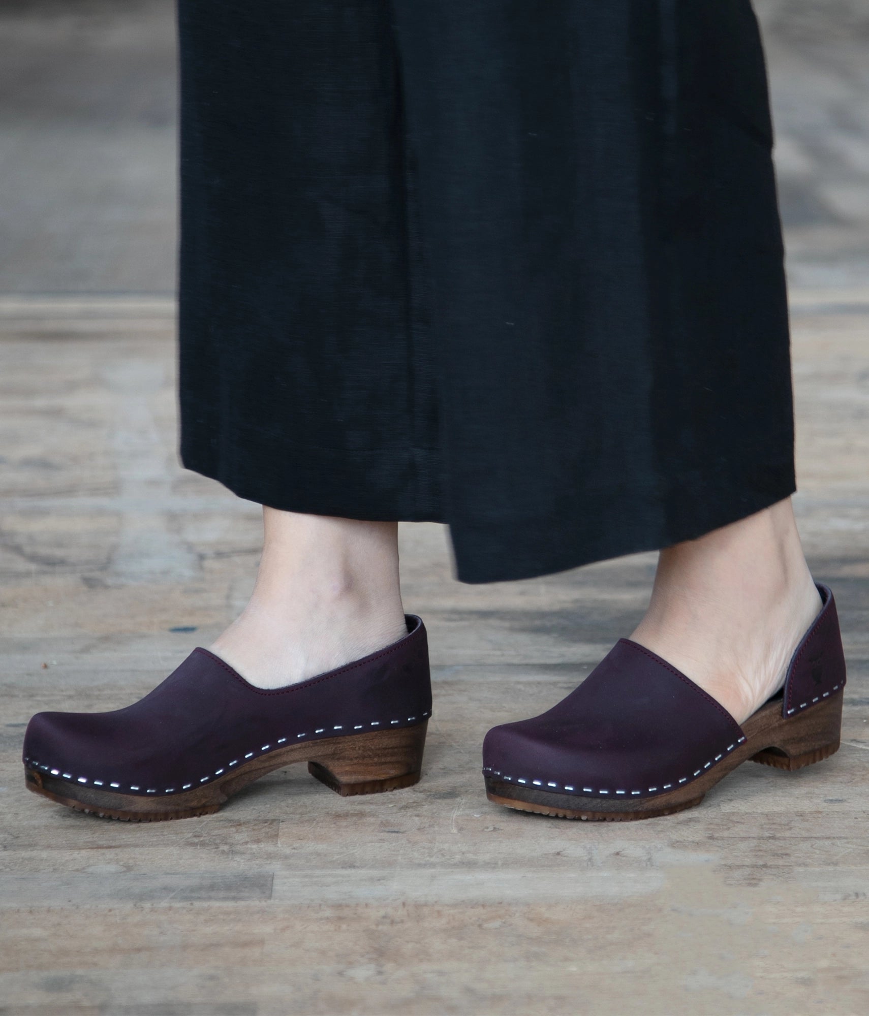 Plum closed-back clogs worn with black pants on a wooden floor. low heeled closed-back clogs in dark purple nubuck leather stapled on a dark wooden base