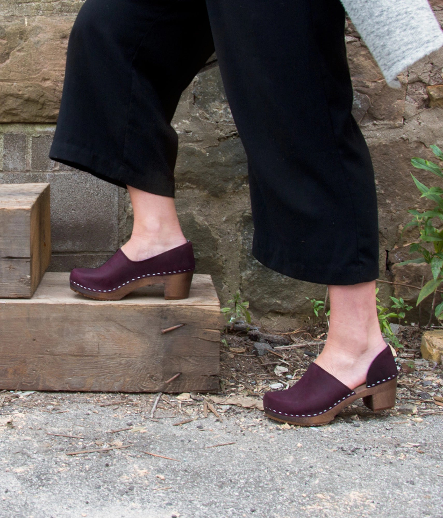 Person wearing plum colored clogs on a stone step with a natural background. high heeled closed-back clogs in purple plum nubuck leather stapled on a dark wooden base