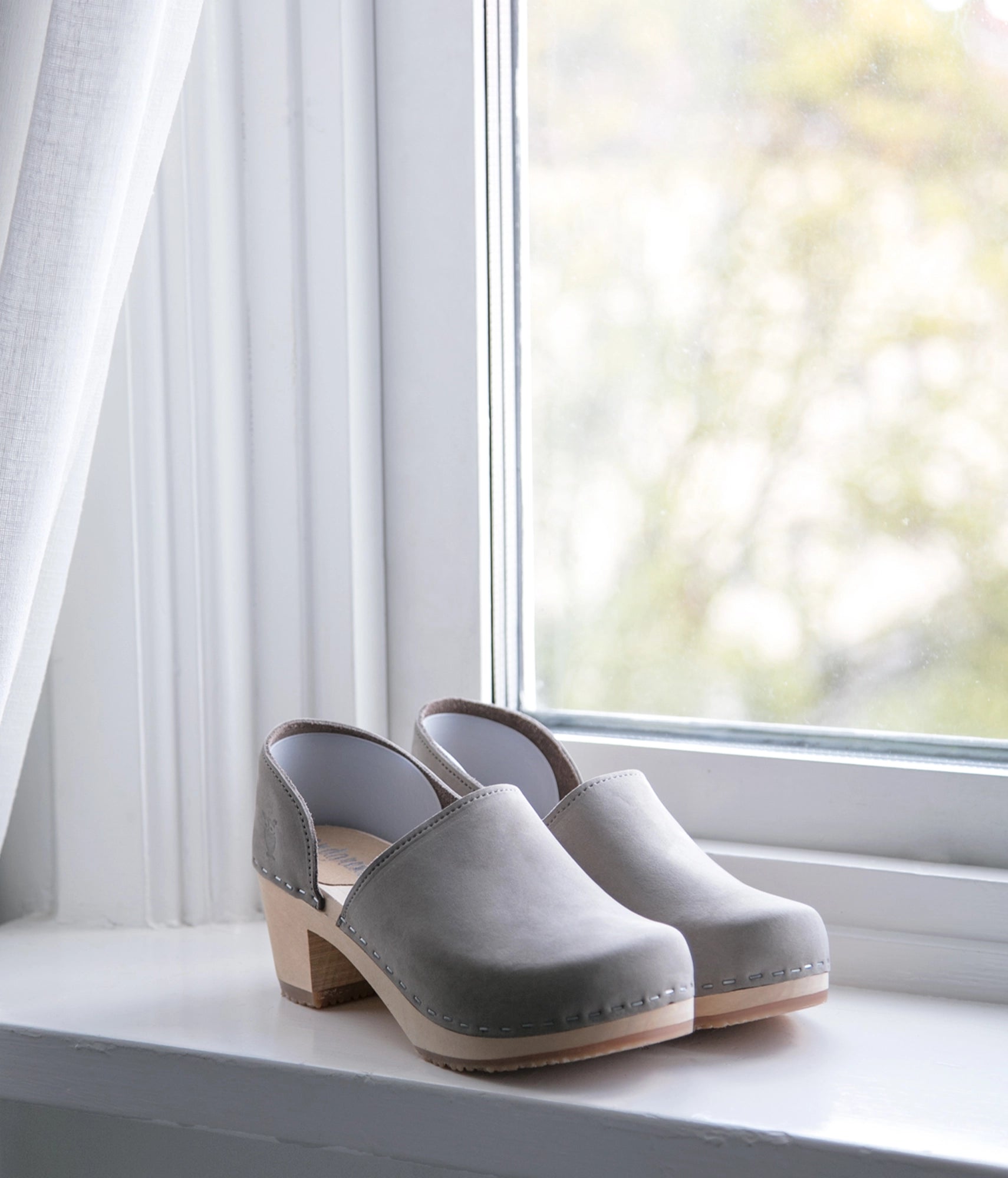 Pair of Stone colored clogs on a windowsill with white curtains and natural light. high heeled closed-back clogs in stone grey nubuck leather stapled on a light wooden base