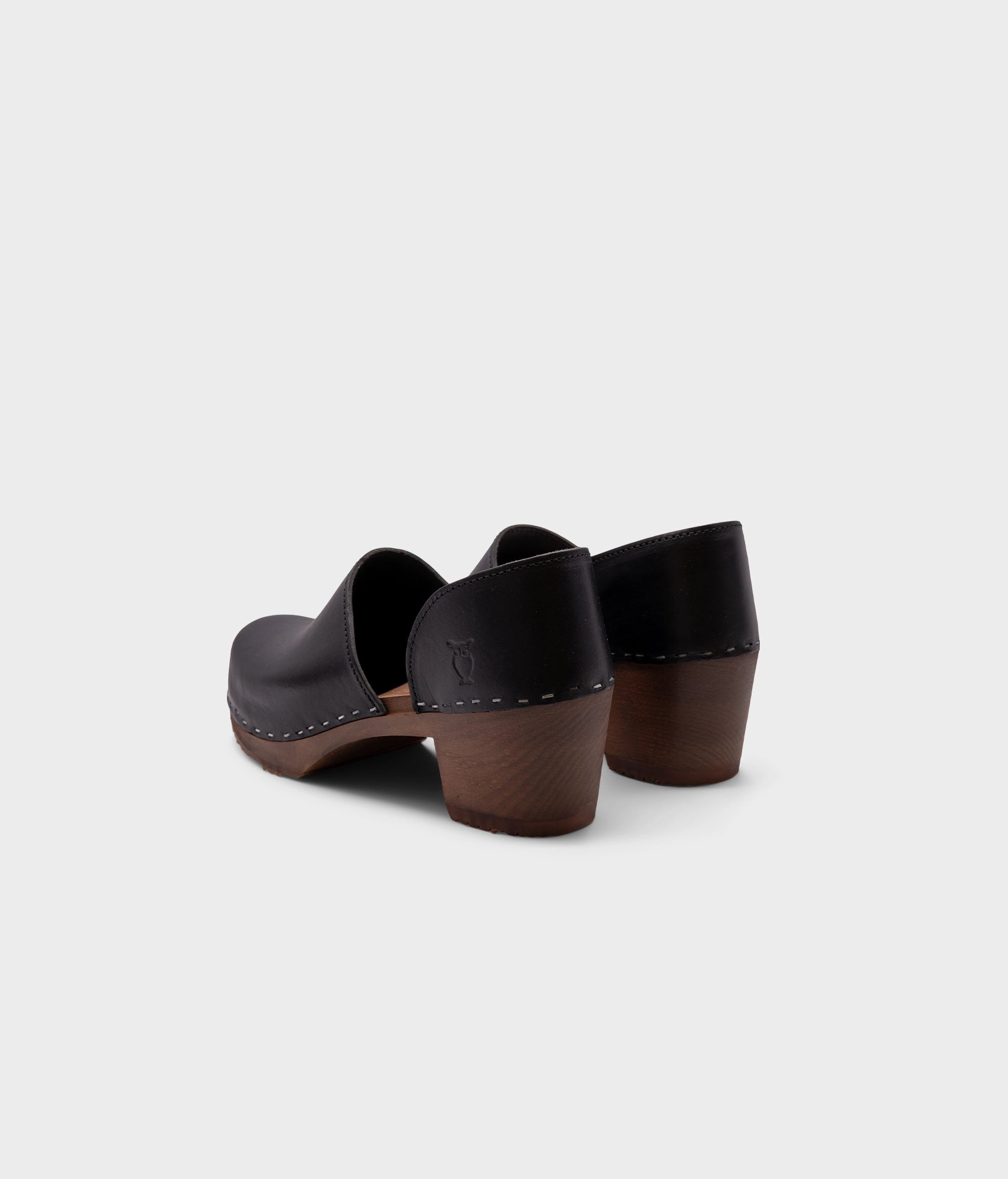 high heeled closed-back clogs in black vegetable tanned leather stapled on a dark wooden base