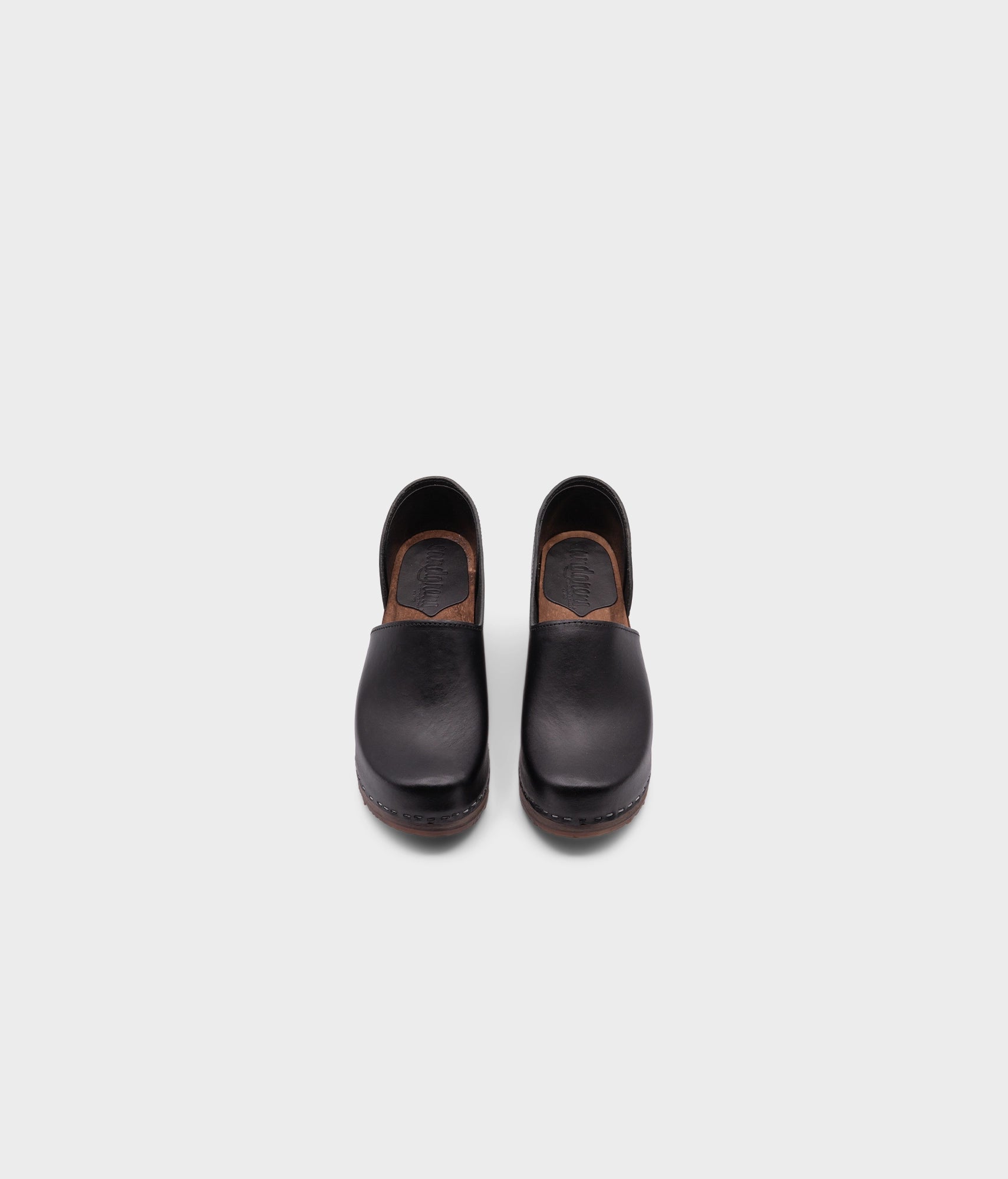 high heeled closed-back clogs in black vegetable tanned leather stapled on a dark wooden base