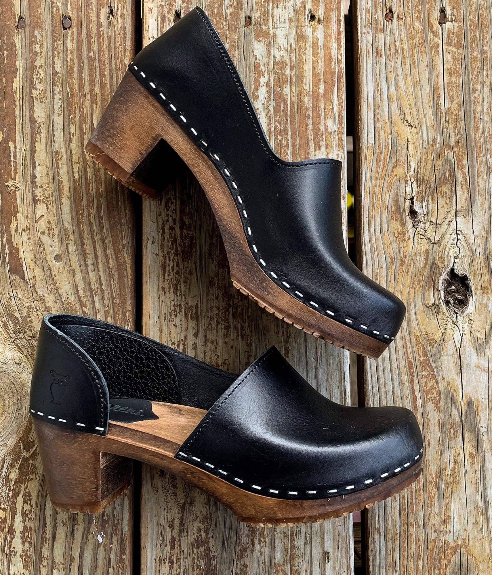 Black leather clogs with wooden soles on a rustic wooden background. High heeled closed-back clogs in black vegetable tanned leather stapled on a dark wooden base