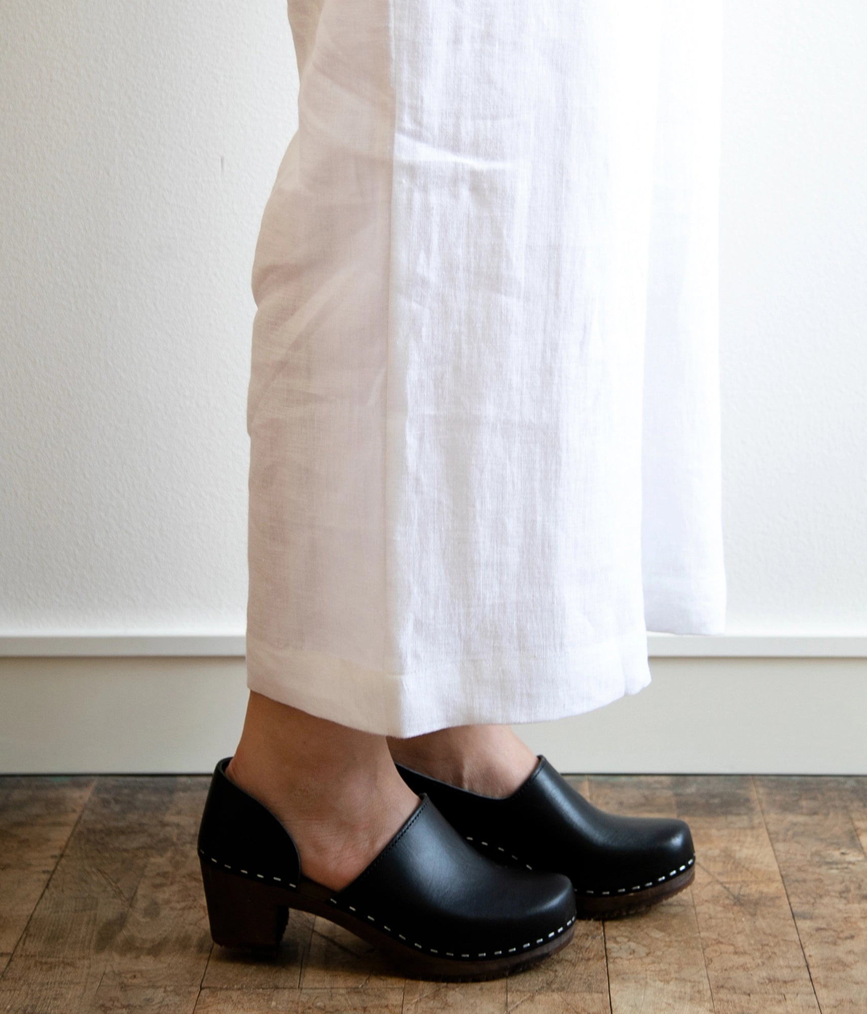 Person wearing black clogs and white pants standing on a wooden floor. High heeled closed-back clogs in black vegetable tanned leather stapled on a dark wooden base