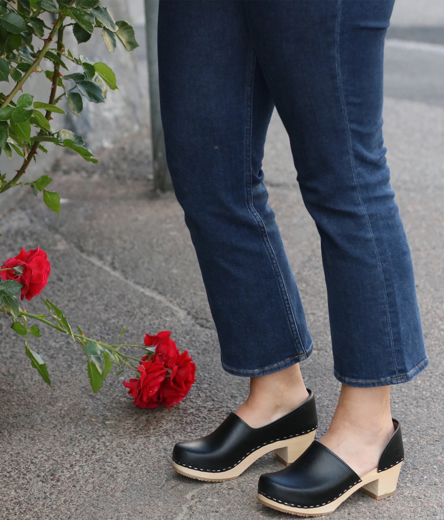 Person wearing blue jeans and black clogs with red flowers on a stone surface. high heeled closed-back clogs in black vegetable tanned leather stapled on a light wooden base