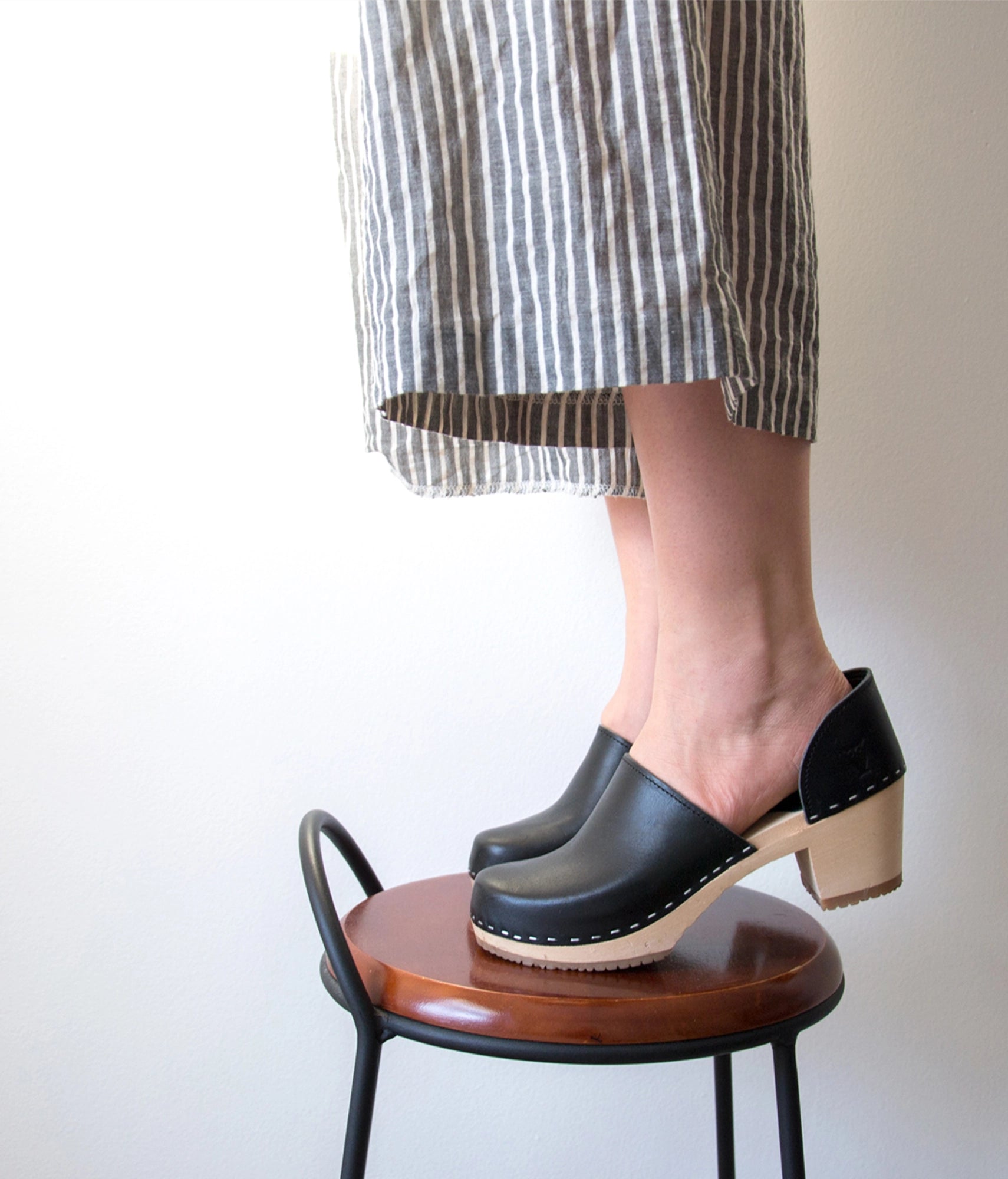 Person wearing black clogs on a wooden stool with a neutral background. high heeled closed-back clogs in black vegetable tanned leather stapled on a light wooden base