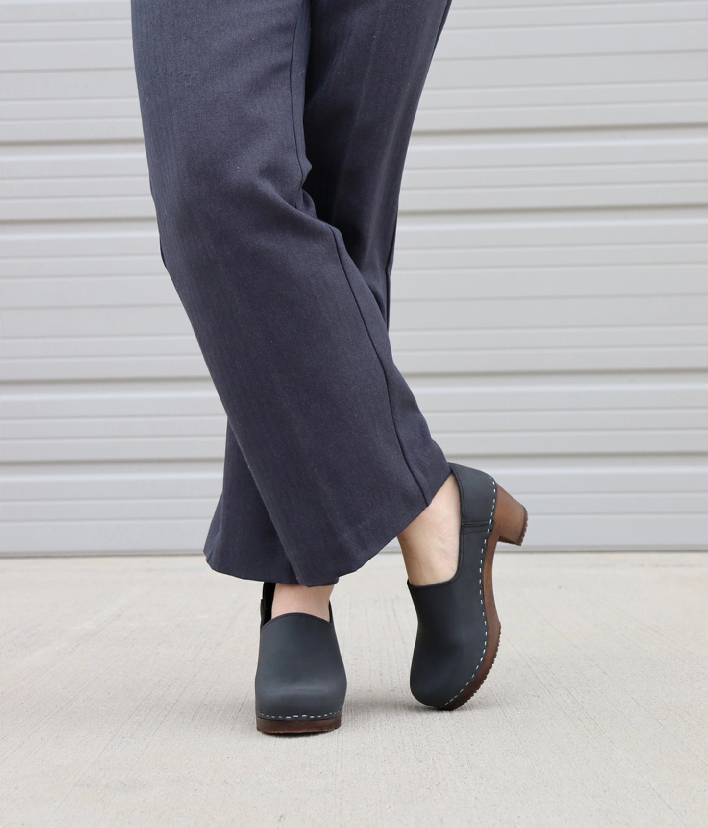 high heeled closed-back clogs in black nubuck leather stapled on a dark wooden base