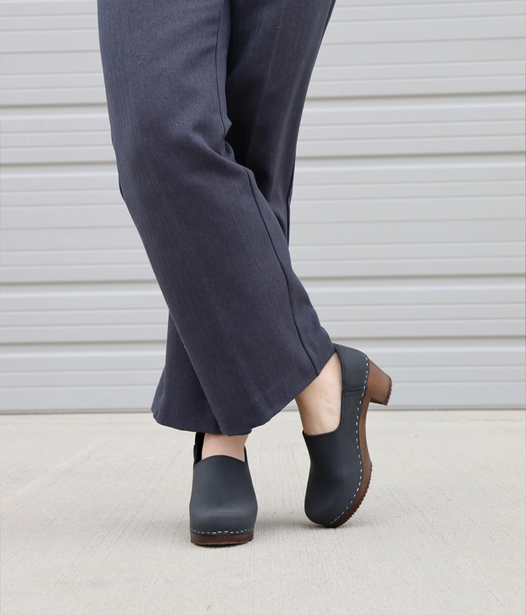 high heeled closed-back clogs in black nubuck leather stapled on a dark wooden base