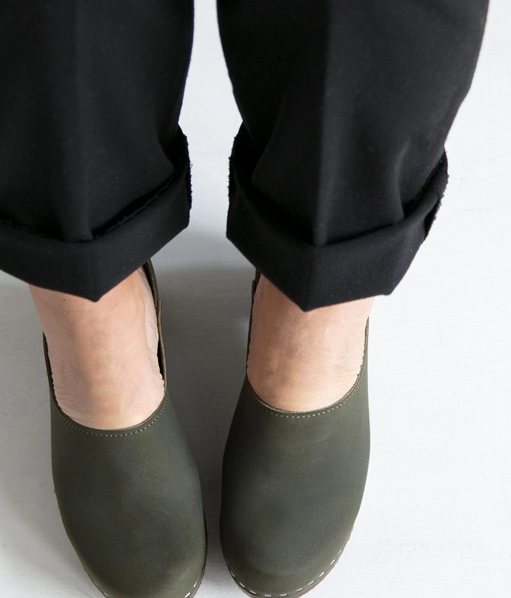 Person wearing dark olive green clogs with black pants on a white background. high heeled closed-back clogs in olive green nubuck leather stapled on a dark wooden base