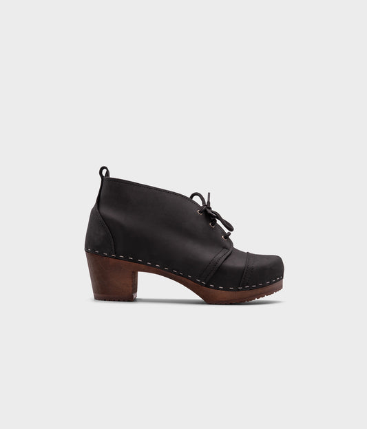 high heeled chukka clog boots in black nubuck leather stapled on a dark wooden base with black laces